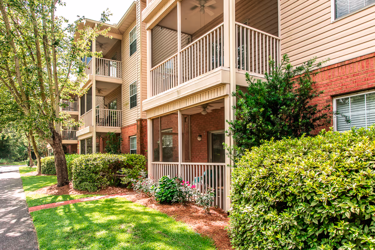 Exterior view of a multi-story residential building with screened porches and balconies, brick and siding facade, and landscaped shrubs and lawn.