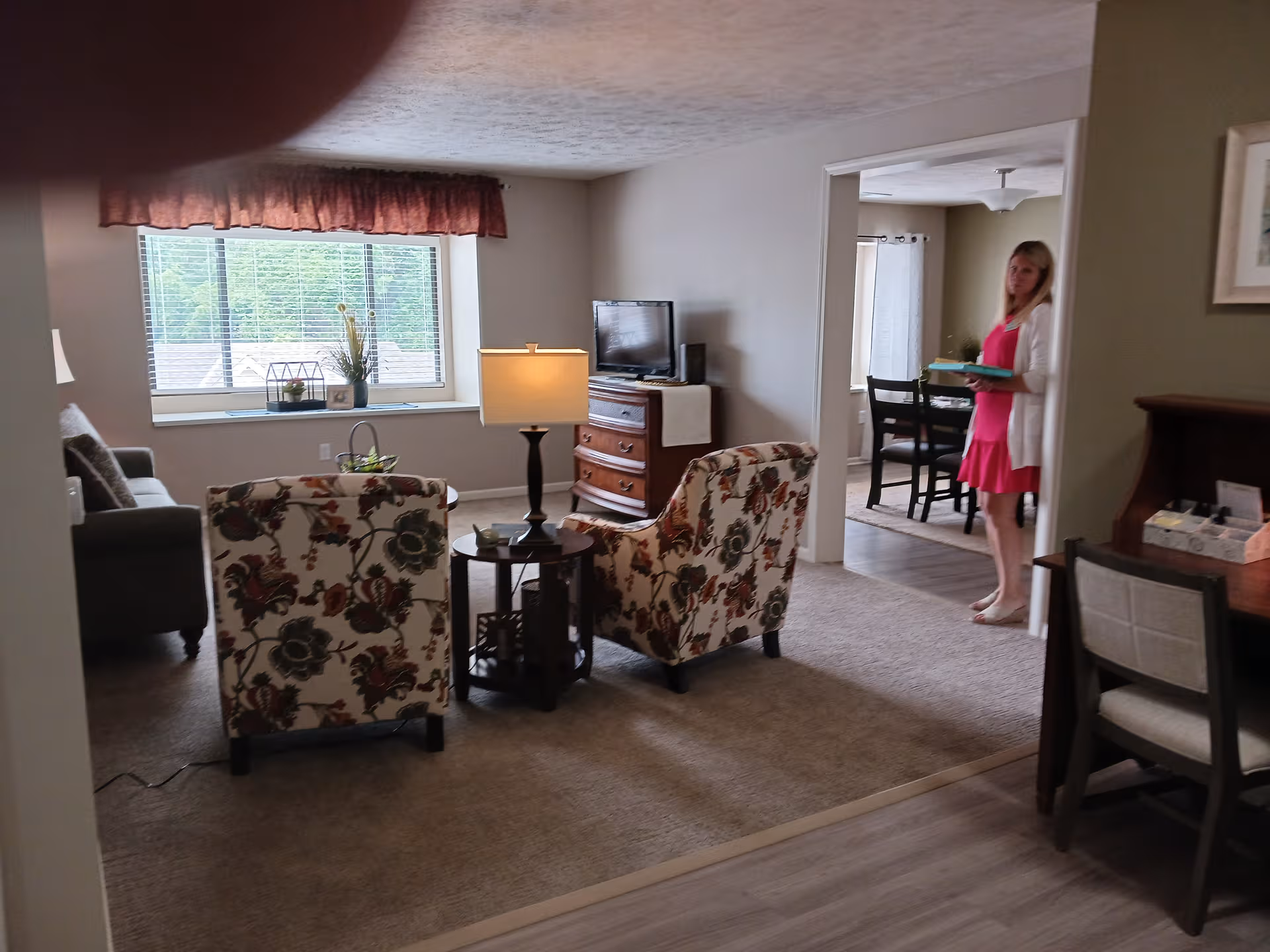 A living room with floral chairs, a lamp, and a woman standing in the doorway.