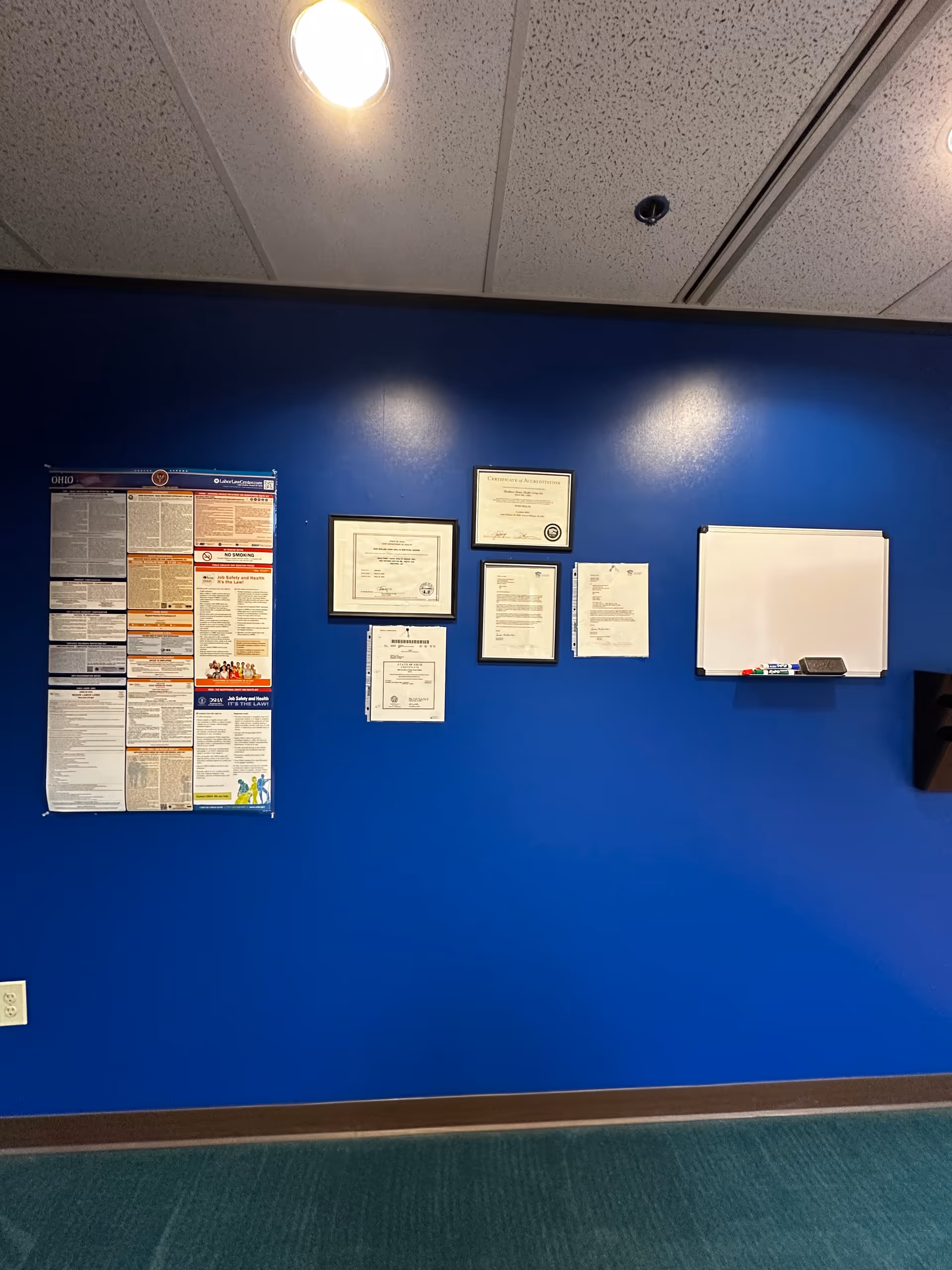 Blue wall with framed certificates, a whiteboard, and a bulletin board indoors.