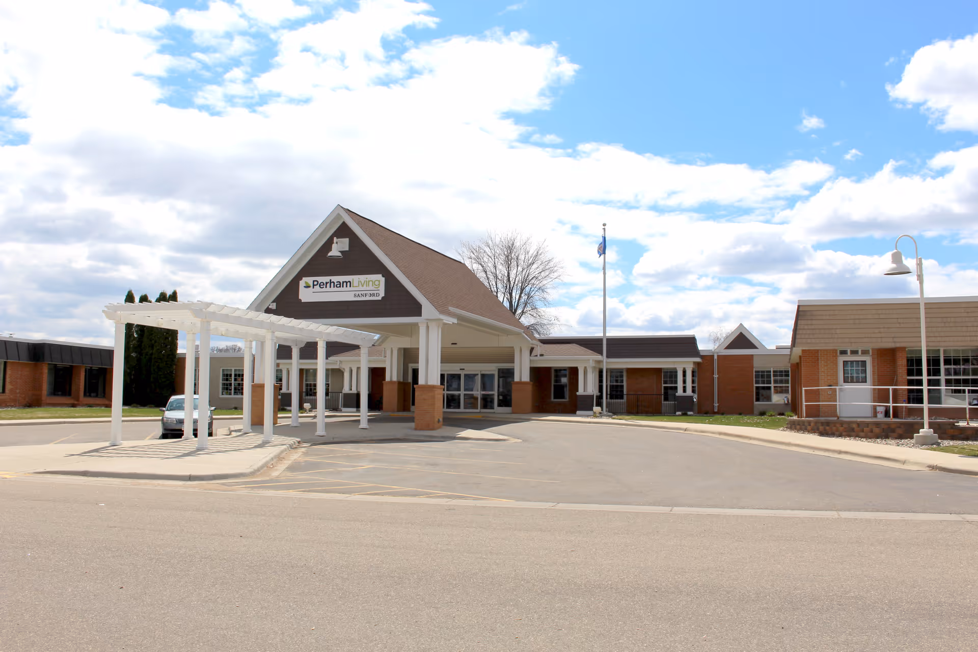 Exterior view of Perham Living facility showing the main entrance with a covered drop-off area, a flagpole, and a clear blue sky with some clouds.