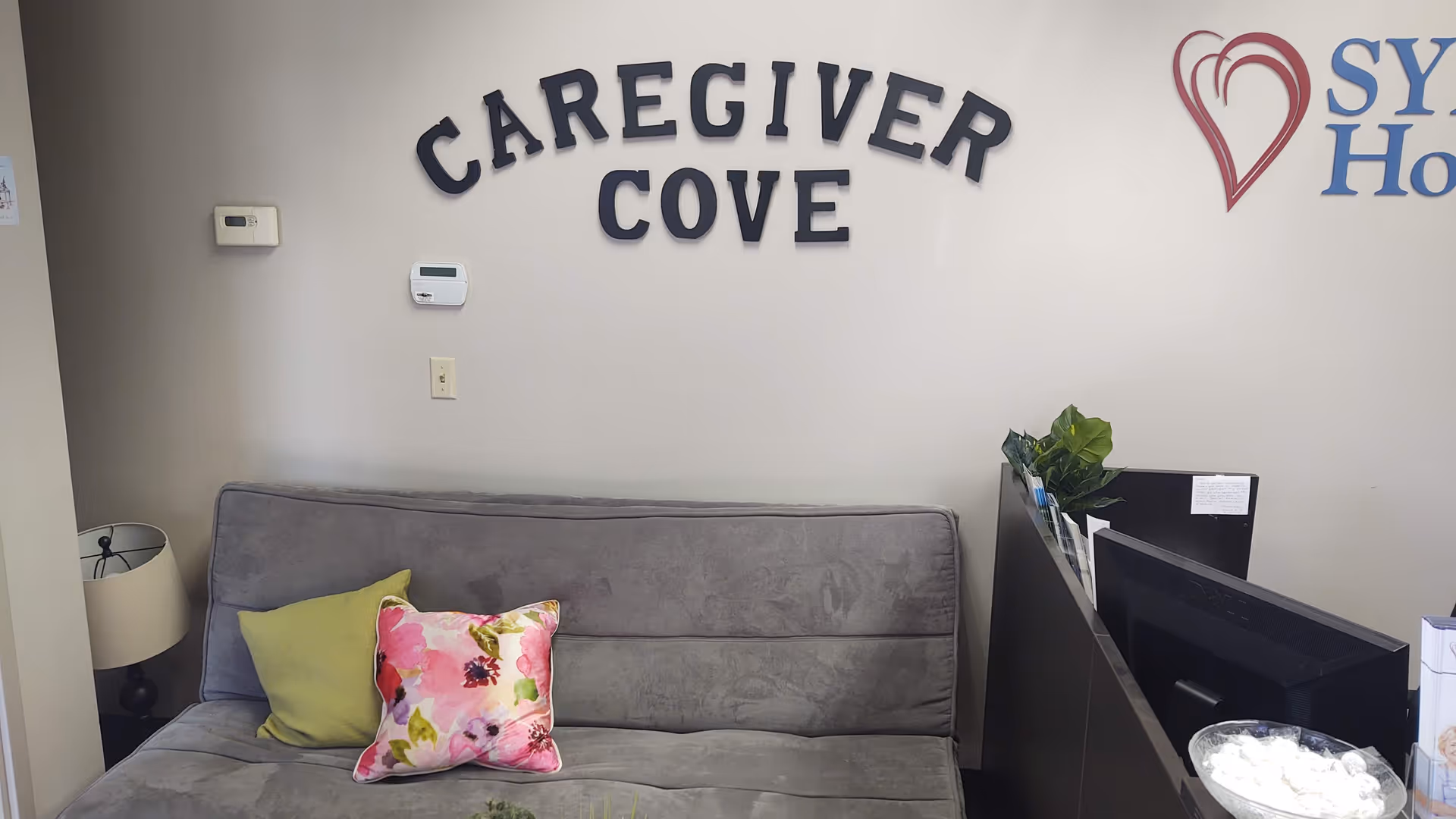 Reception area with gray sofa, colorful pillows, plant, and front desk signage reading Caregiver Cove.