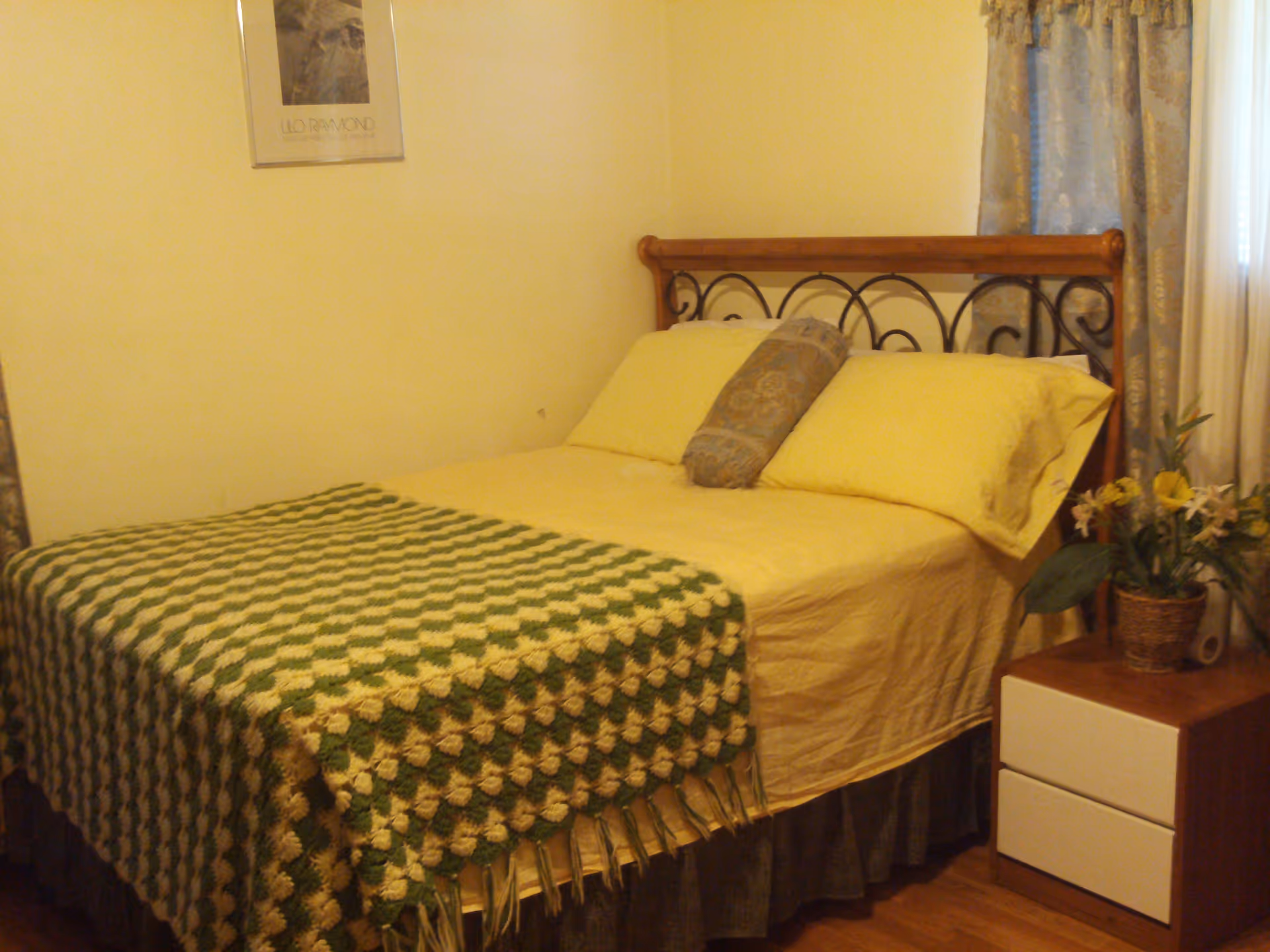 A cozy bedroom with a bed featuring a wooden and metal headboard, yellow bedding, a decorative pillow, and a green and yellow crocheted blanket. Next to the bed is a wooden nightstand with two white drawers and a potted plant with flowers. A window with patterned curtains is partially visible on the right side, and a framed picture hangs on the wall above the bed.