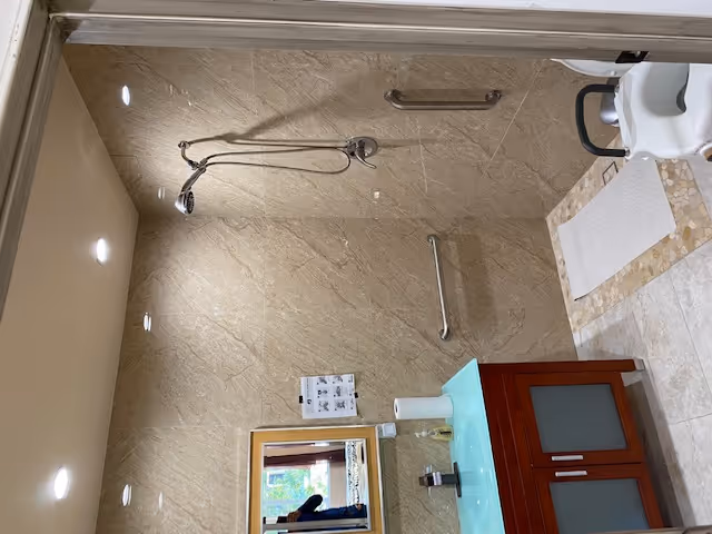 A bathroom with beige marble walls and floor tiles. There is a walk-in shower area with a handheld showerhead and two grab bars on the walls. A white bath mat is placed on the shower floor. To the left, there is a wooden vanity with a frosted glass countertop, a soap dispenser, and a paper towel roll. A mirror is mounted above the vanity, reflecting a window and part of a person taking the photo.