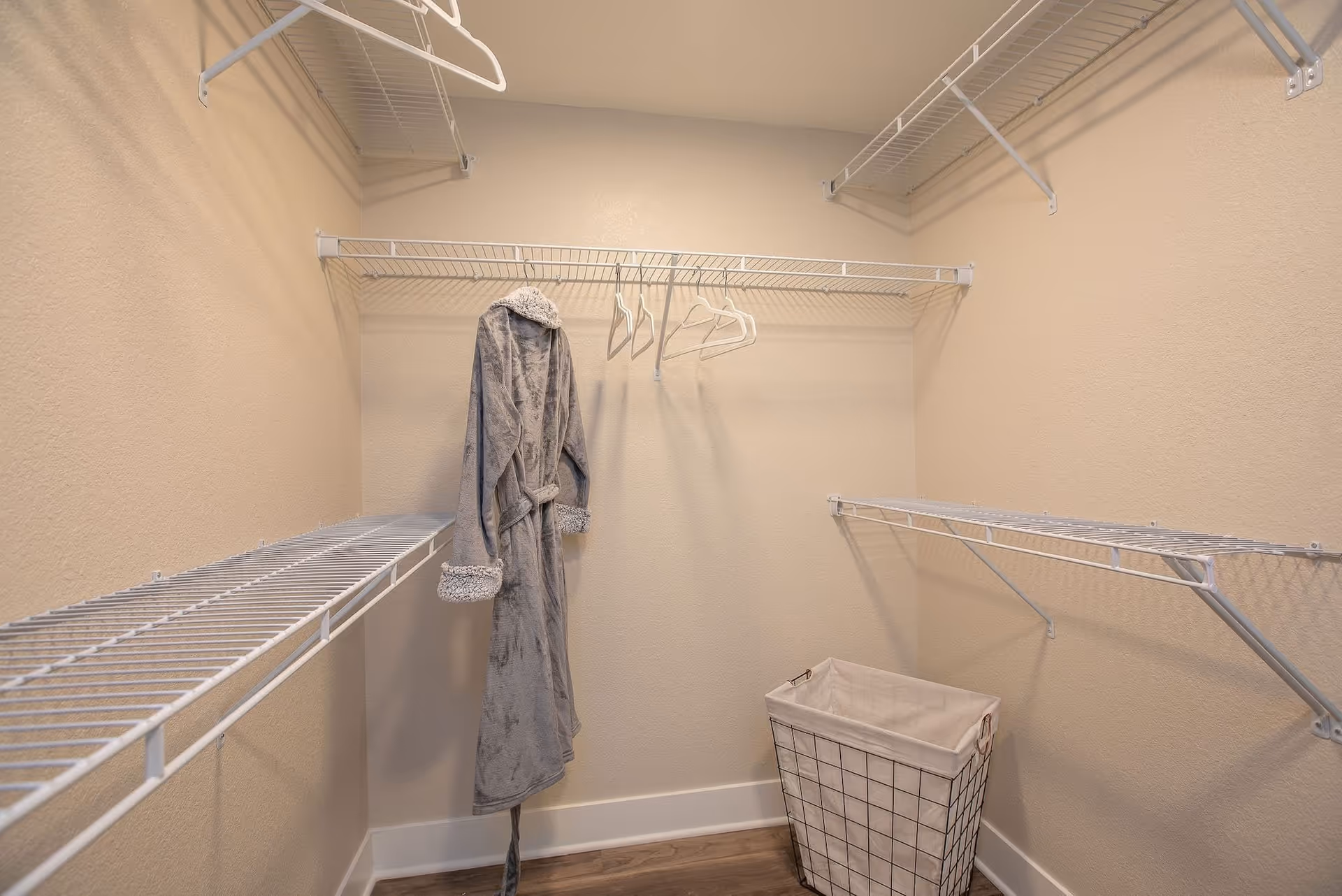 A walk-in closet with beige walls and white wire shelving on three sides. A gray robe hangs on one of the shelves along with several empty white hangers. A wire laundry basket with a fabric liner is placed on the wooden floor in the corner.