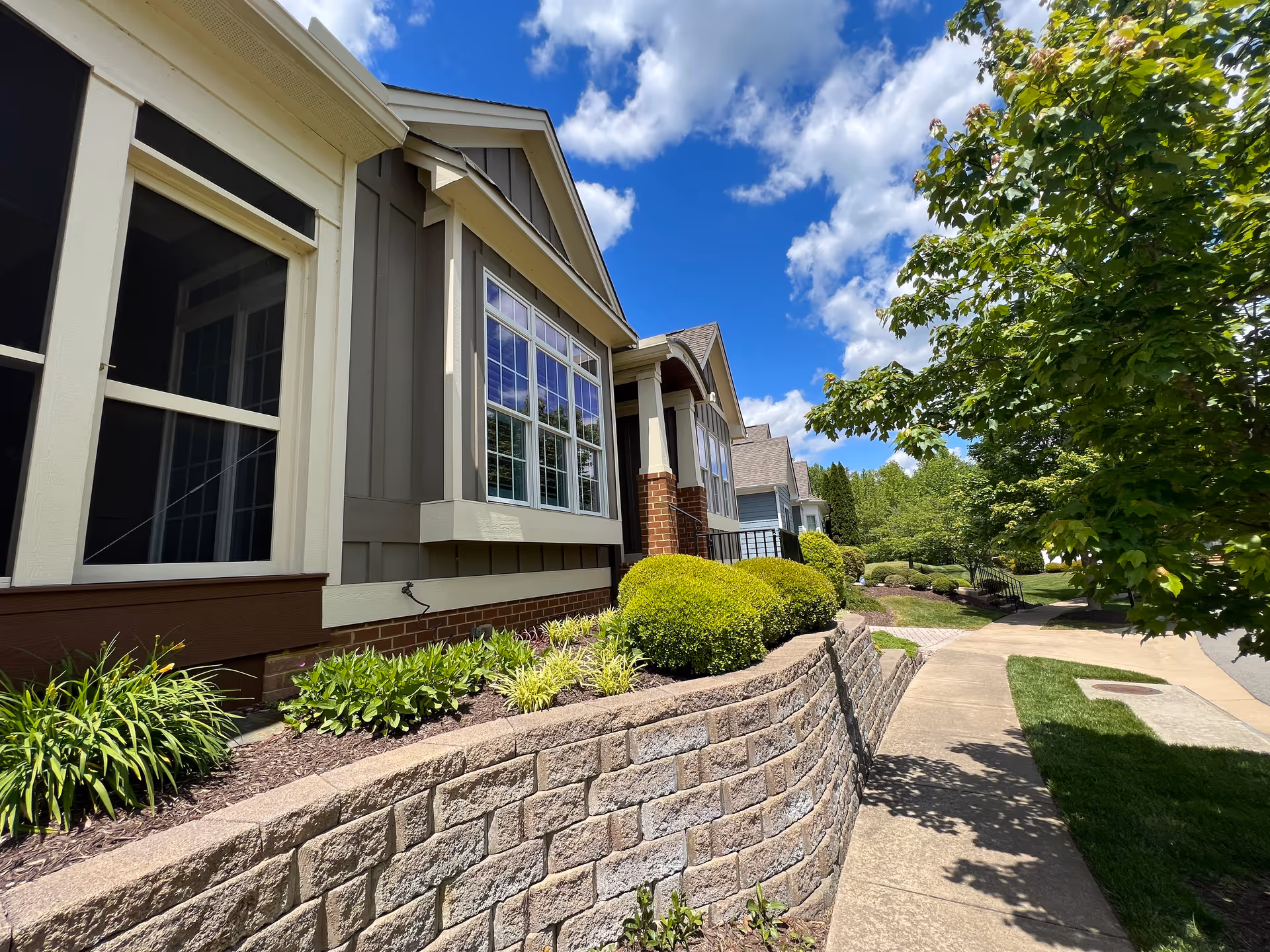 Exterior view of a senior living facility building with beige and brown siding, large windows, and a brick retaining wall with neatly trimmed bushes and plants. A sidewalk curves alongside the building under a partly cloudy blue sky with green trees nearby.