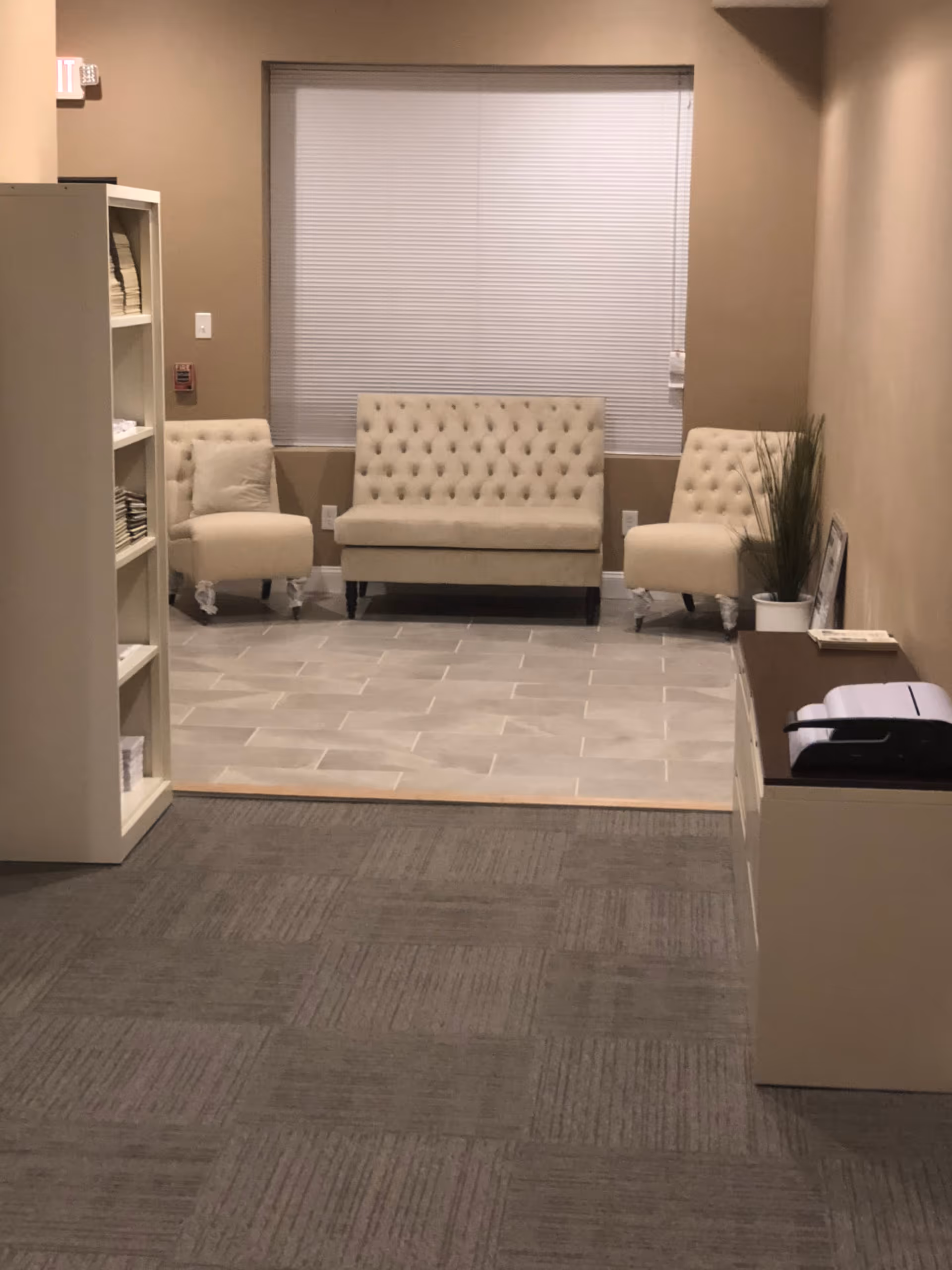 A neutral, modern waiting area with tufted chairs, shelves, and a plant.