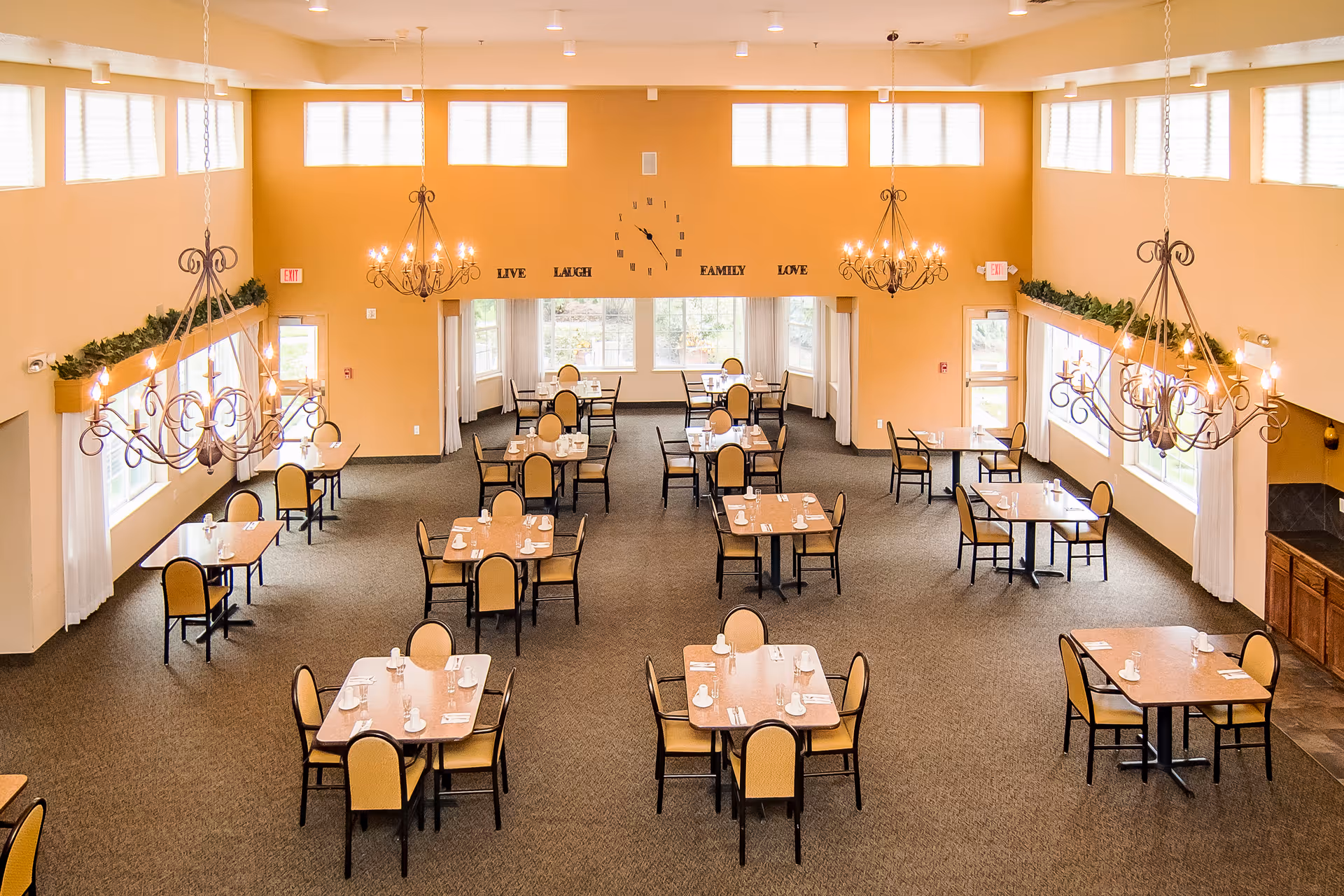 A spacious dining room with multiple tables and chairs arranged neatly. Each table is set with white cups, saucers, and utensils. The room has large windows allowing natural light to fill the space, beige walls, and decorative chandeliers hanging from the ceiling. On the far wall, there is a clock and the words 'LIVE LAUGH FAMILY LOVE' displayed.