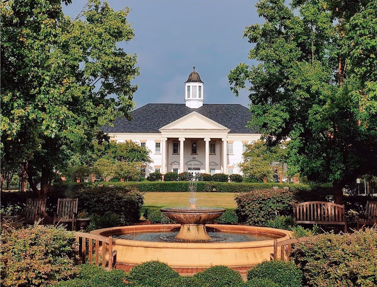 Front view of a white, columned building with a central cupola, fountain, and landscaped grounds.