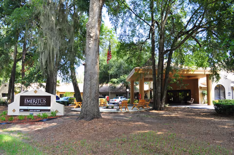 Front entrance of a senior living facility with a covered porte-cochère, outdoor seating, large trees, and a sign reading "Emeritus".