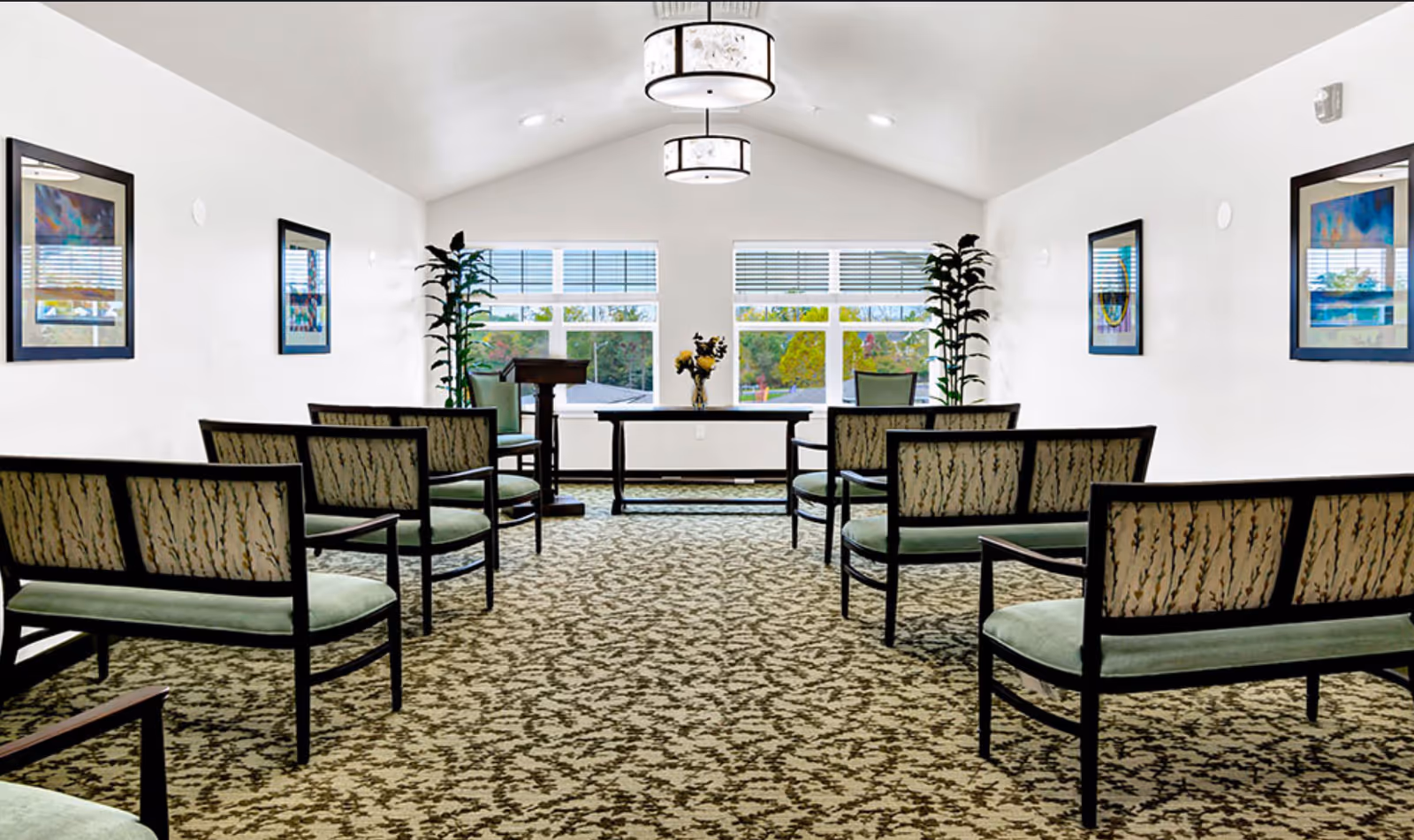 Bright multipurpose meeting room with rows of upholstered benches facing a table and lectern in front of large windows.