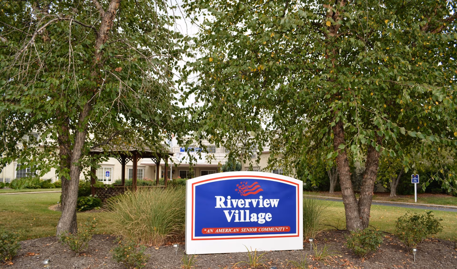 A landscaped entrance sign reading "Riverview Village" sits in front of trees and the facility building.