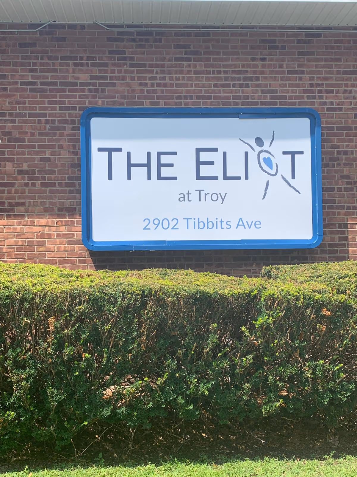 Blue-framed sign on a brick building that reads 'THE ELIOT at Troy 2902 Tibbits Ave' above trimmed hedges.