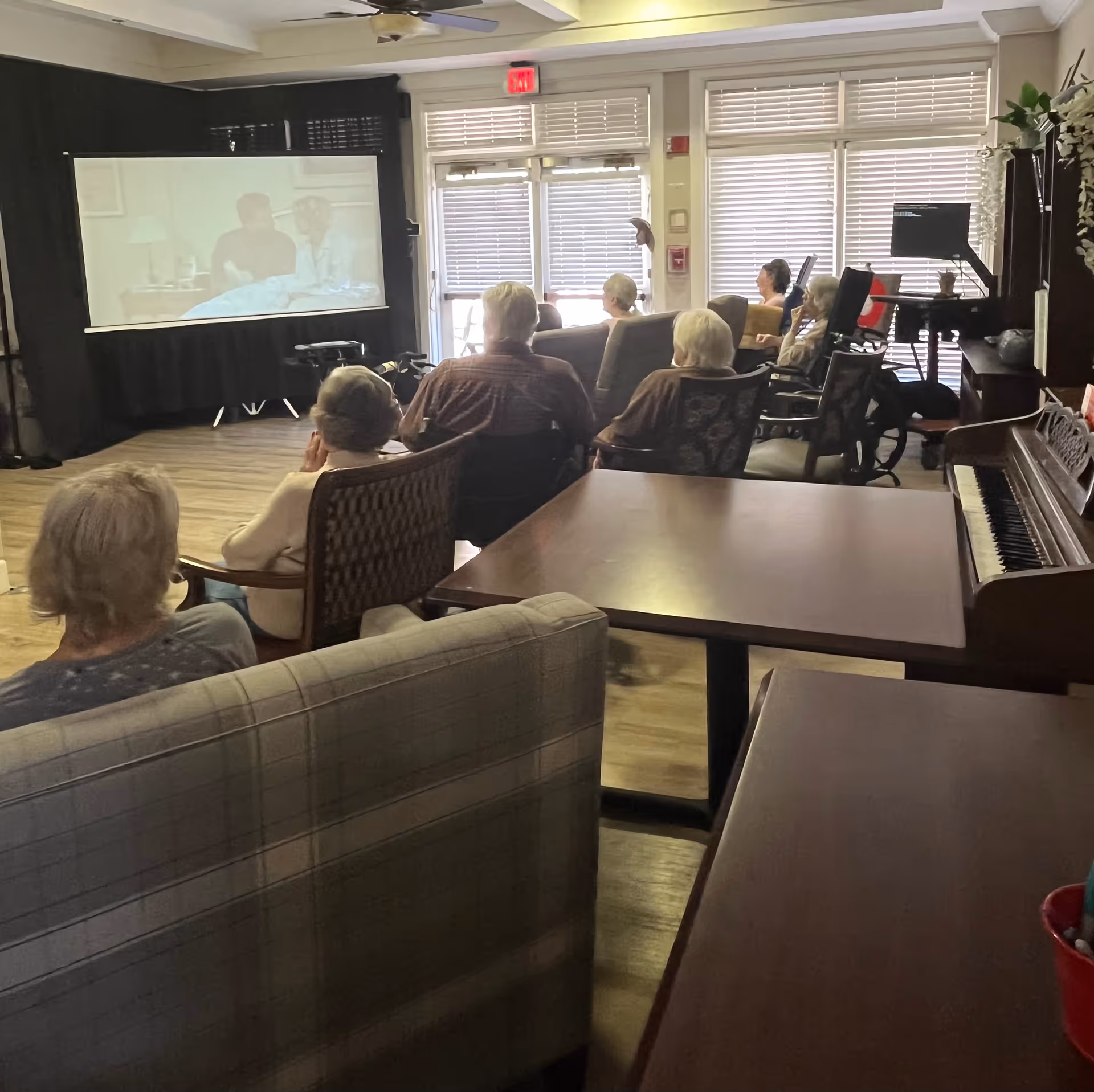 Elderly residents watch a movie in a living room lounge with seating and a piano.