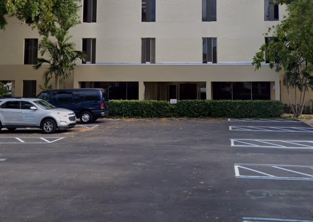 Front exterior of a beige, multi-story senior facility with a parking lot and hedges.