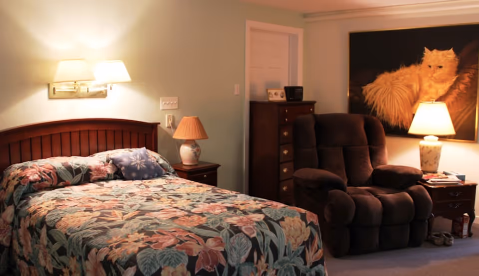 Cozy bedroom with a floral bedspread, bedside lamps, a plush recliner, and framed cat artwork on the wall.