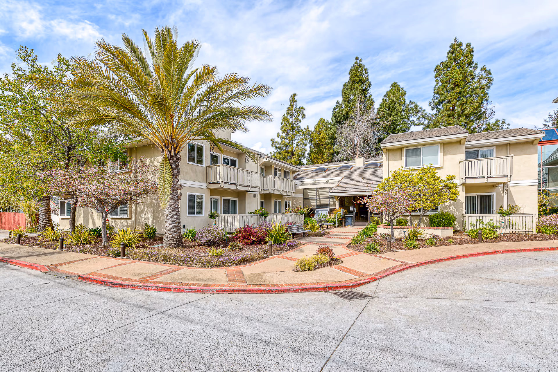 Exterior view of a two-story senior living facility building with beige walls, balconies, and a landscaped entrance featuring palm trees, flowering shrubs, and a paved walkway under a partly cloudy sky.