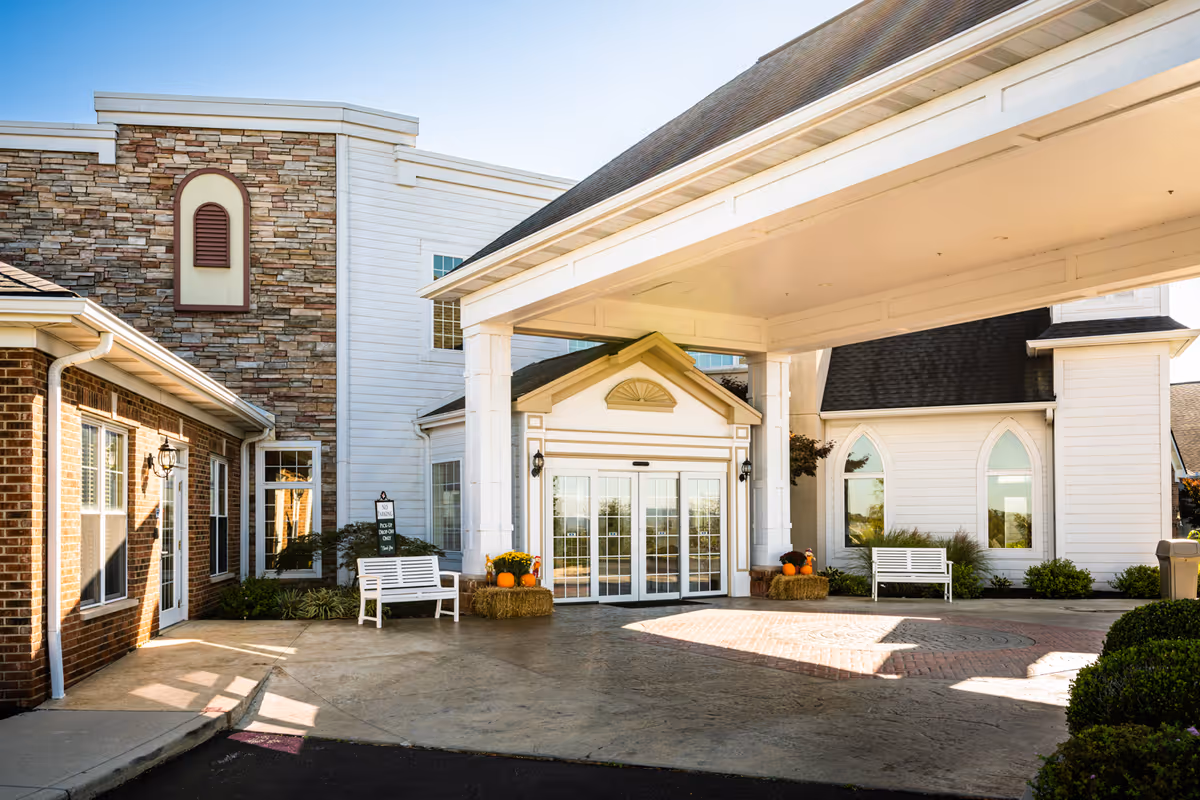 Covered entrance and porte-cochère of a nursing facility with white benches, pumpkins on hay bales, and brick and stone facade.