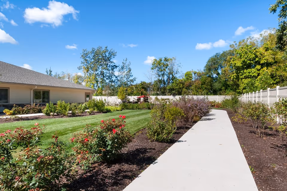 Concrete walkway through a landscaped garden with shrubs and roses beside a single-story building under a blue sky.