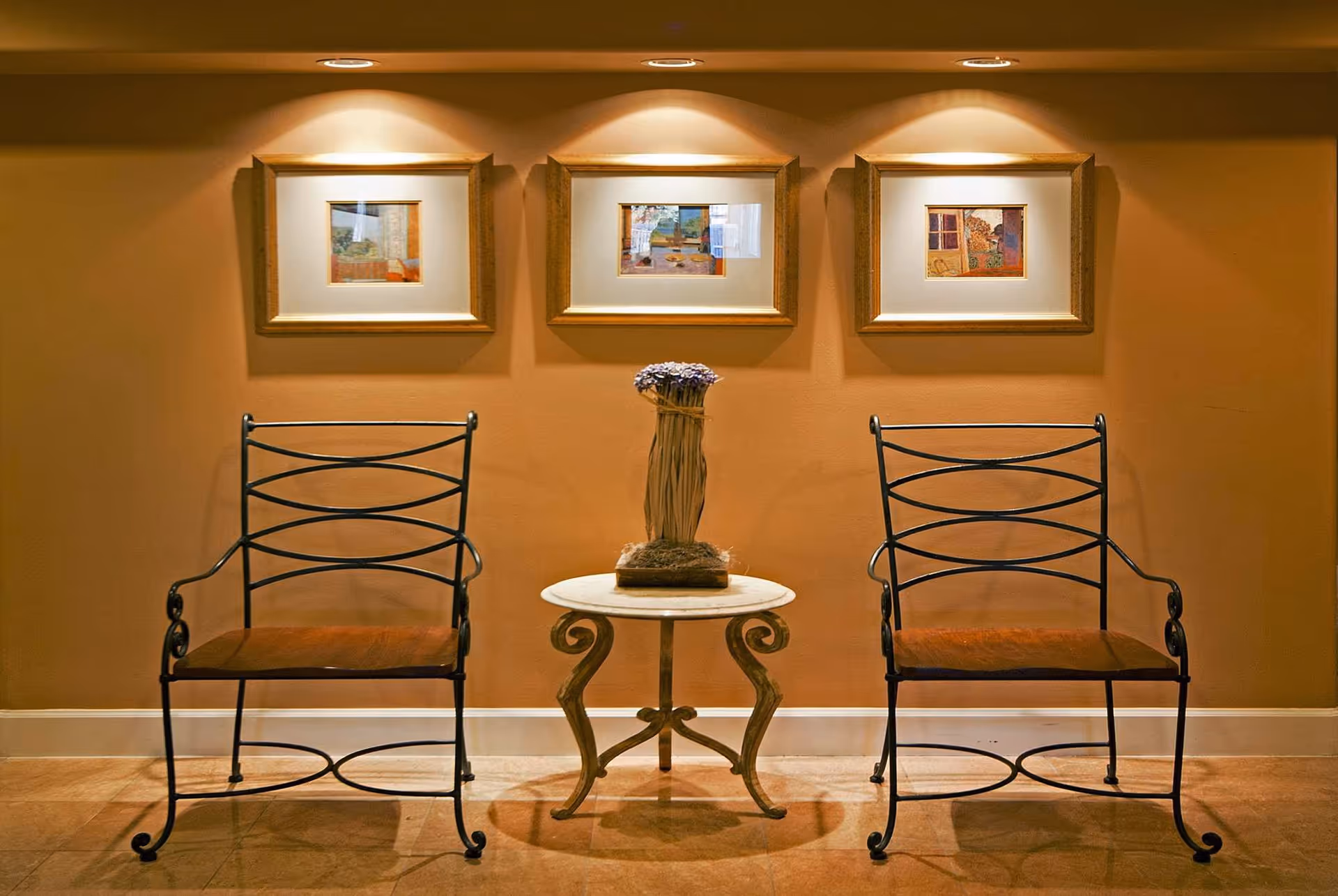 Two wrought iron chairs with wooden seats are positioned on either side of a small round table with ornate legs. On the table is a decorative bundle of dried flowers. Behind the chairs, the wall is painted a warm beige color and features three framed pictures illuminated by individual overhead spotlights.