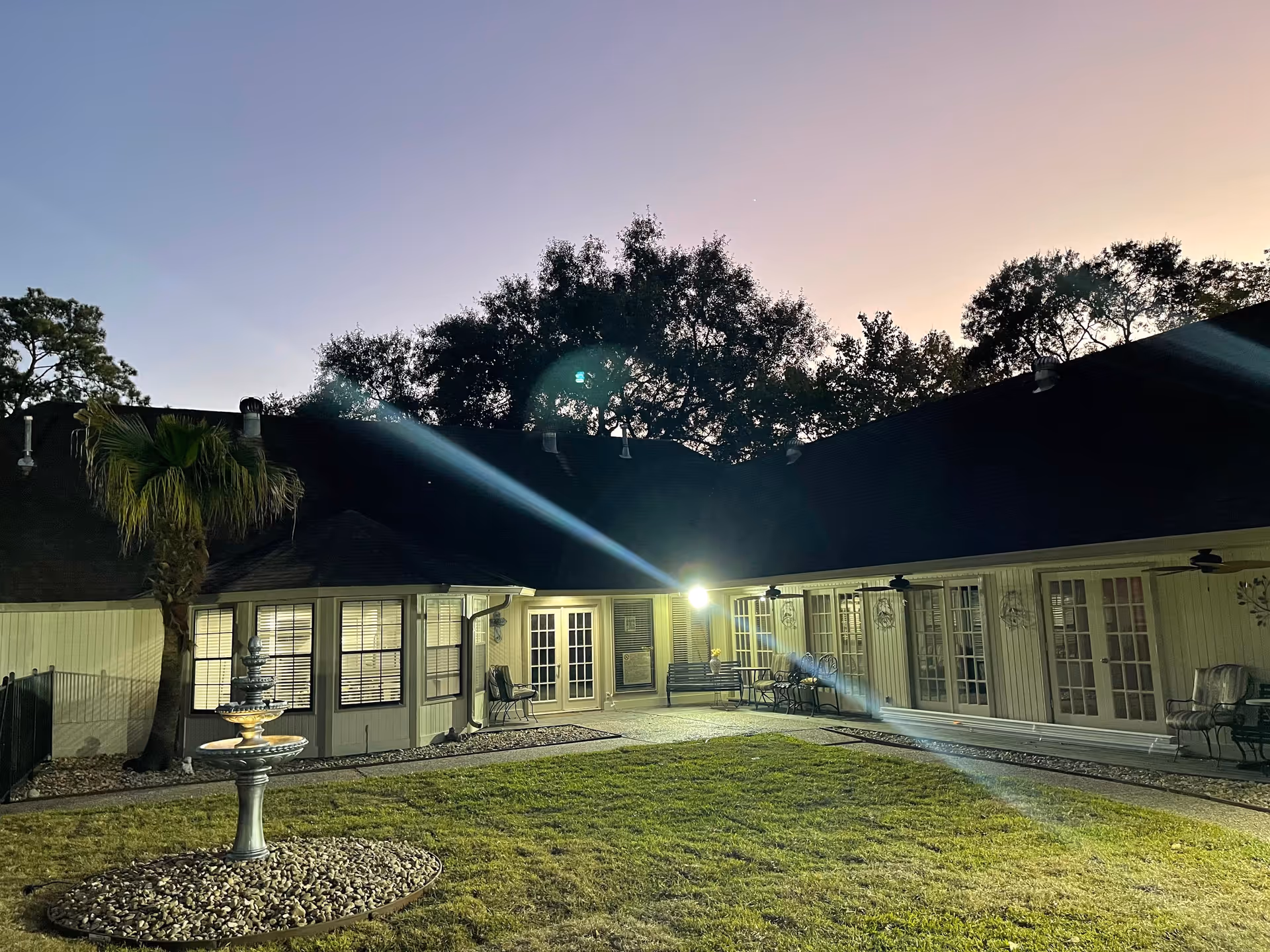 Dusk view of a courtyard with a small fountain and grassy lawn surrounded by a one-story building with lit windows and glass doors.