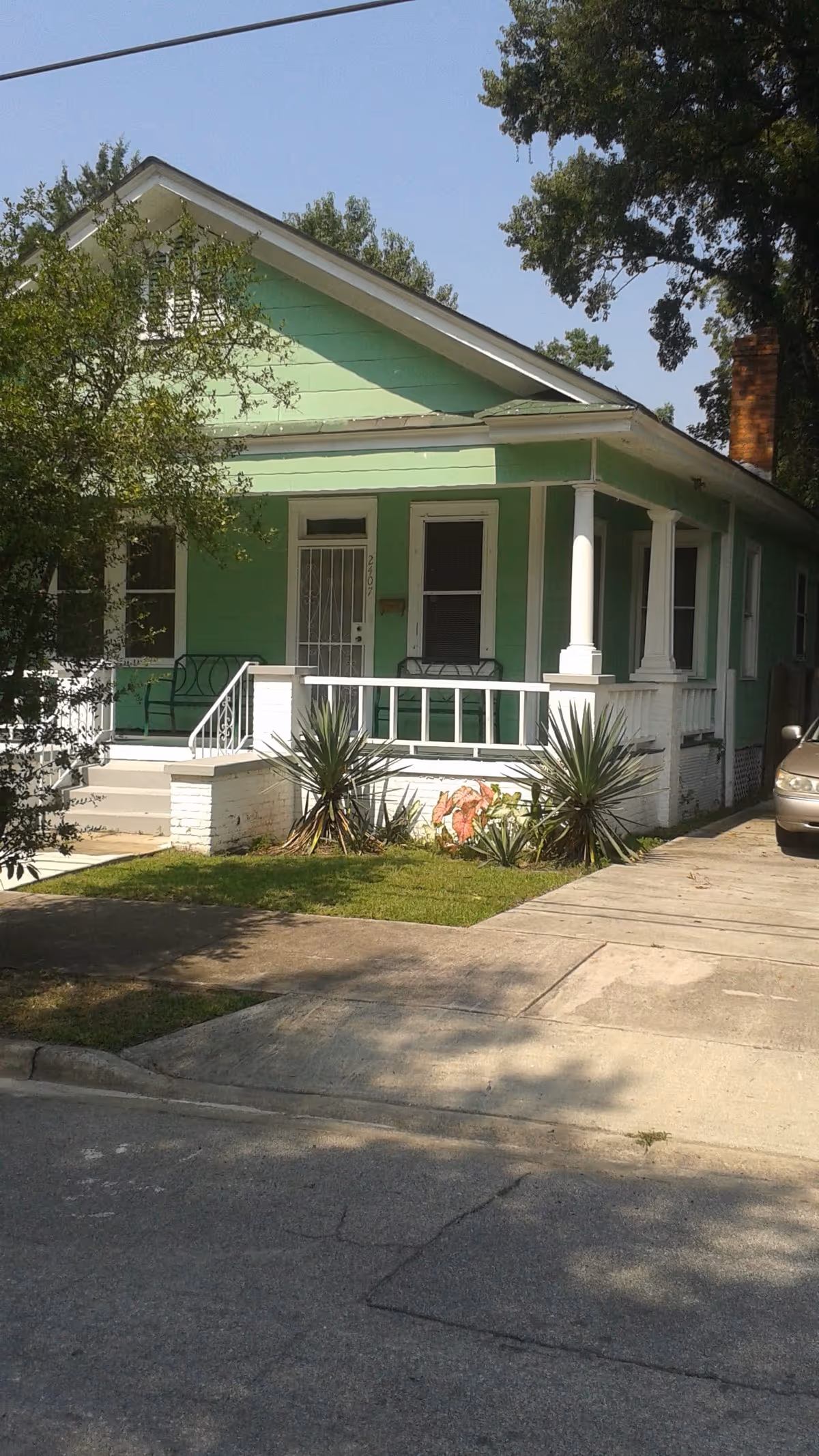 Exterior view of a single-story green house with a white porch railing and columns. There are plants and small bushes in front of the porch, and a car is partially visible parked on the driveway to the right. The sky is clear and blue.