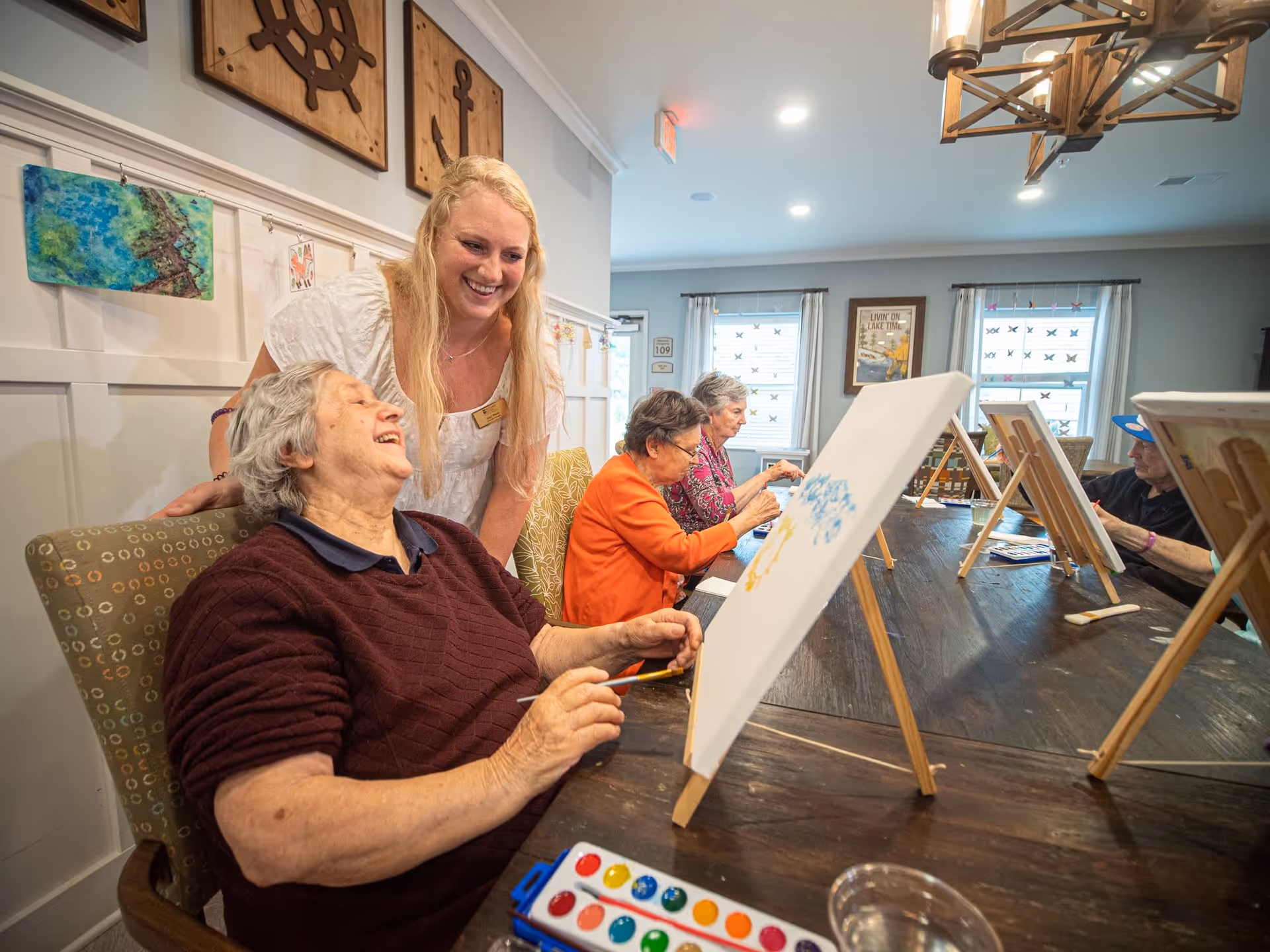 A group of elderly women seated at a long wooden table engaged in painting on canvases set on easels. A smiling caregiver or staff member stands behind one of the women, interacting warmly. The room is decorated with nautical-themed wall art and has large windows with curtains letting in natural light.