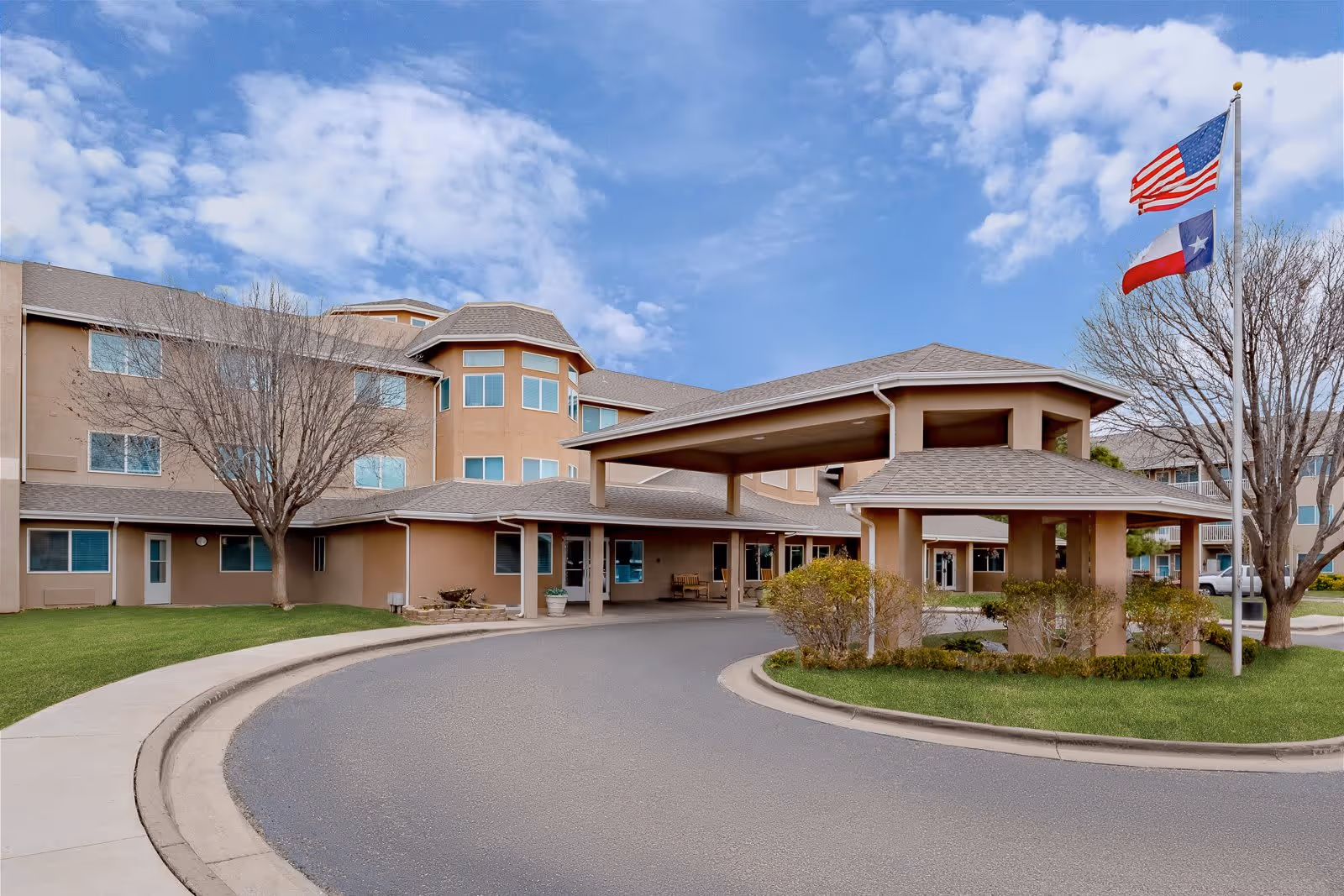 Exterior view of Ventura Place - Sky Active Living facility showing a three-story building with beige walls and multiple windows. There is a covered driveway entrance with a small landscaped area featuring bushes and two flagpoles flying the American and Texas flags. The sky is partly cloudy.
