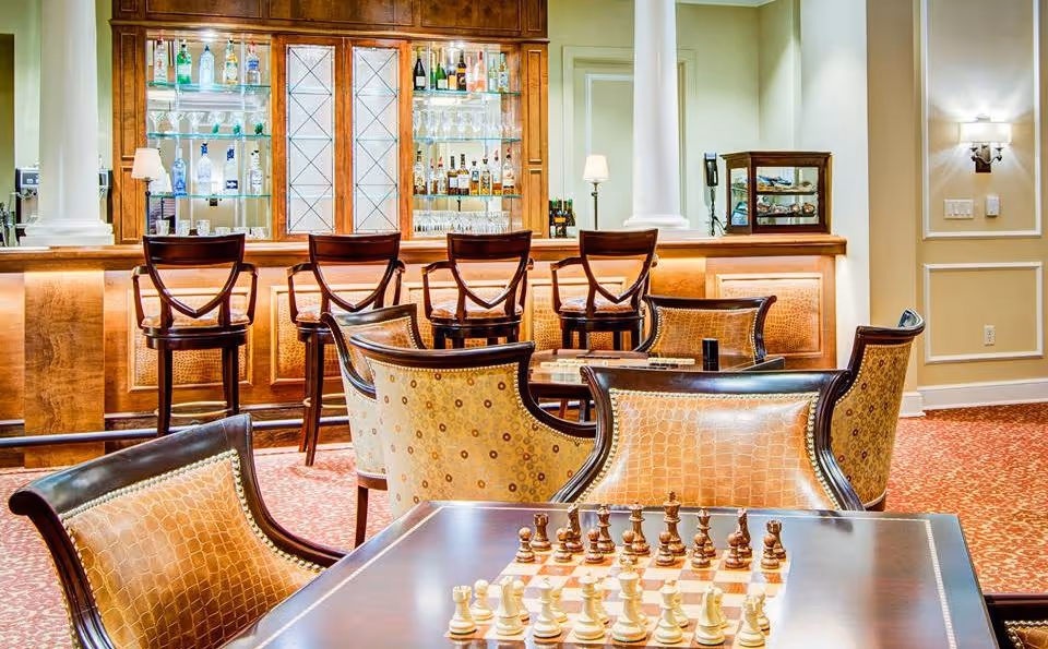 Elegant indoor lounge and bar area with a chessboard on a table in the foreground, upholstered chairs, and bar stools in front of a backlit cabinet of bottles.
