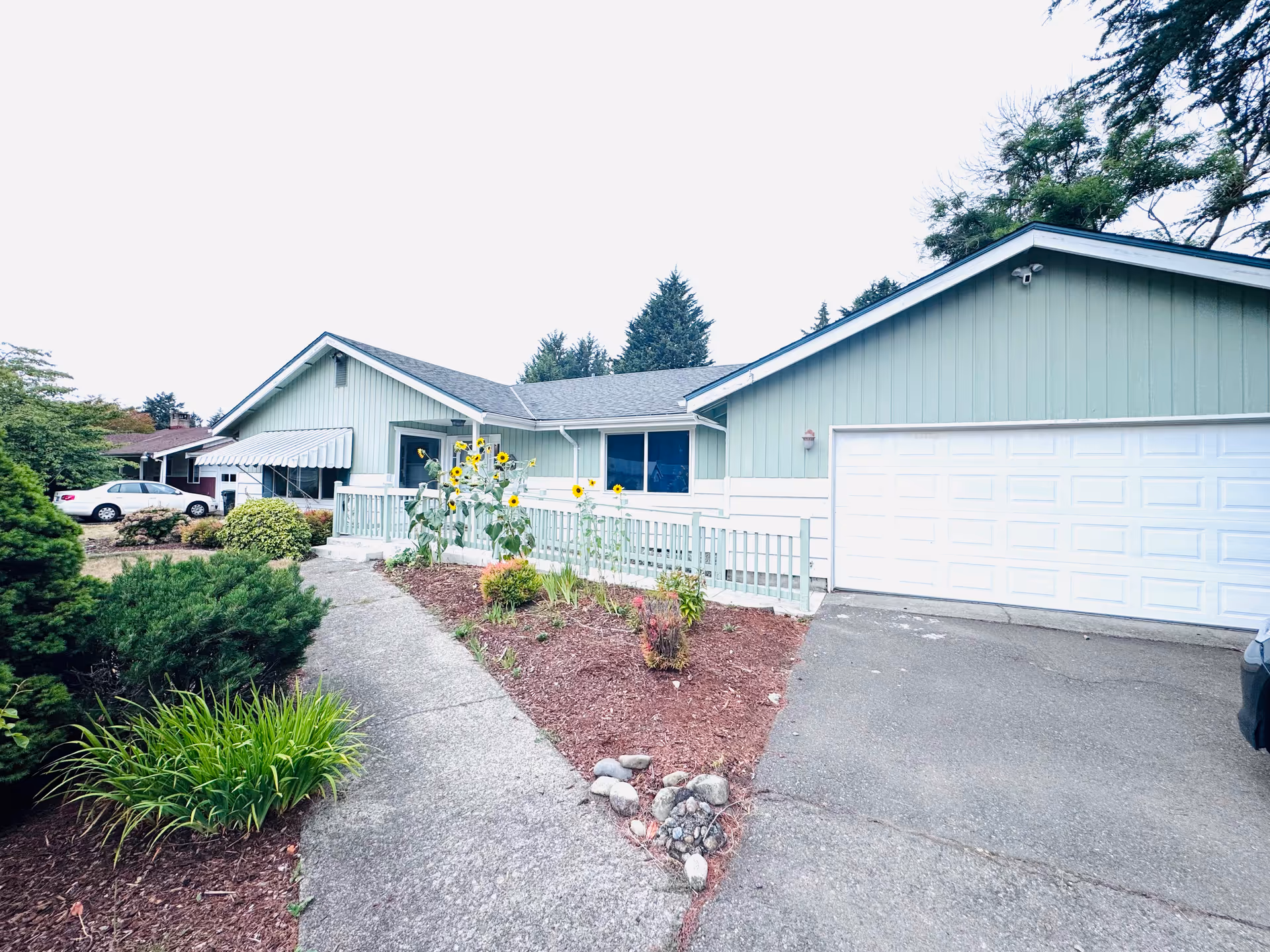 Single-story pastel green home with a two-car garage, walkway, and garden beds.