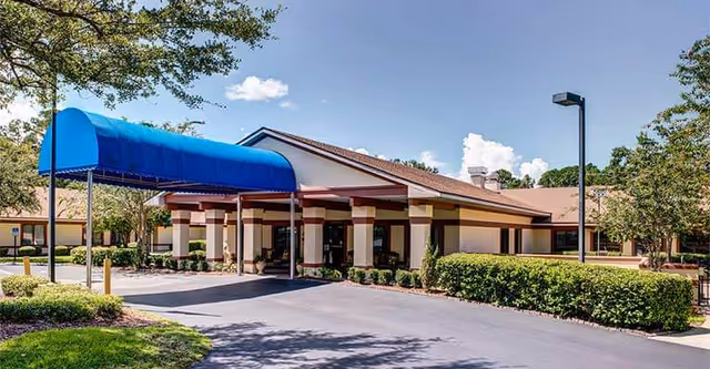 Senior living facility entrance with blue canopy, driveway, and landscaped surroundings.