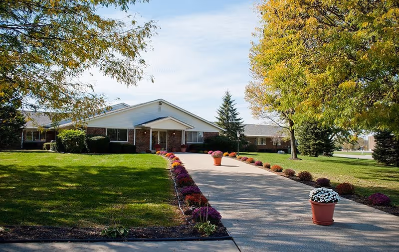 A single-story brick building with a white gabled roof, surrounded by green lawns and trees with autumn foliage. A paved driveway lined with colorful flower pots leads up to the entrance of the building under a small covered porch.