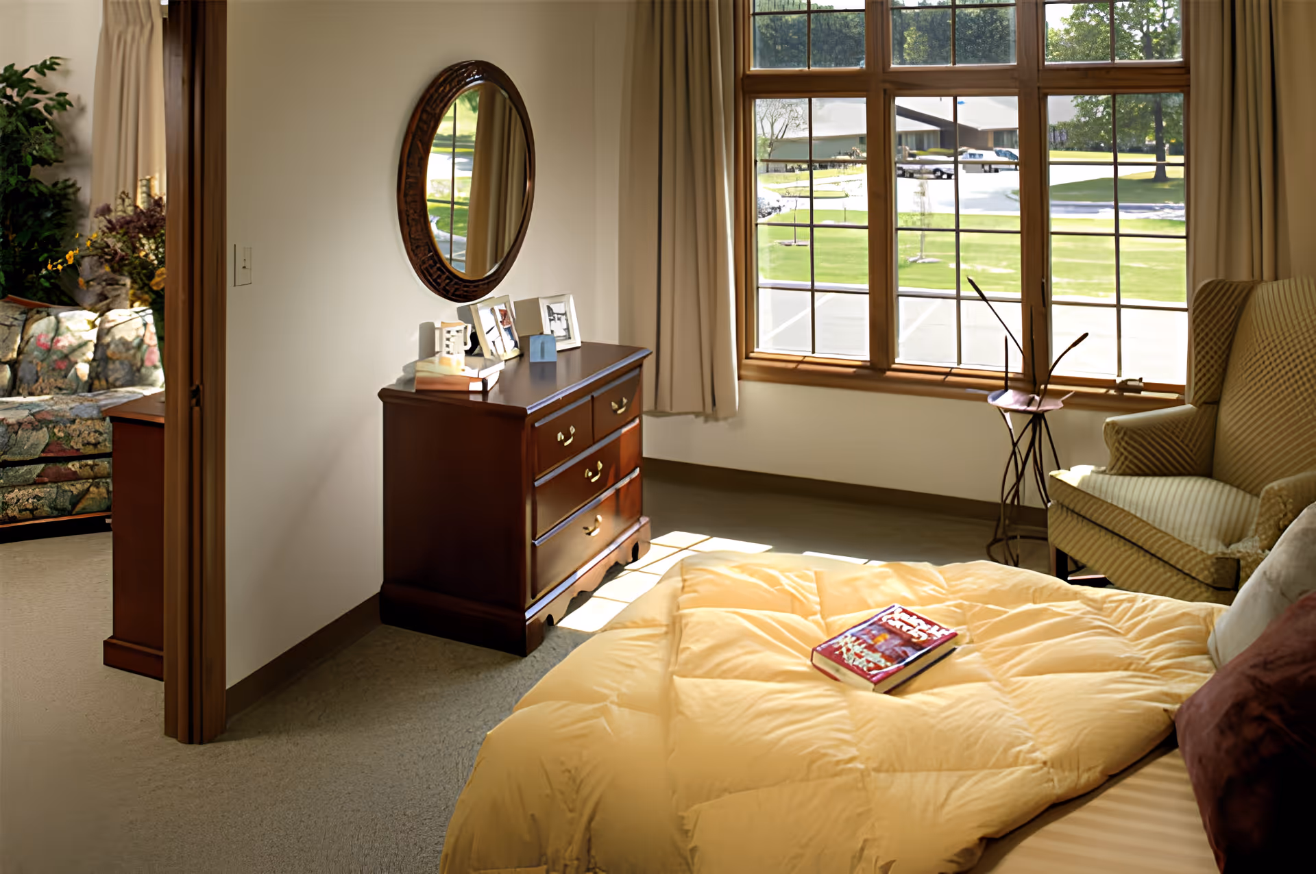 Sunlit bedroom with a bed, wooden dresser topped with photos and a round mirror, an armchair, and large windows overlooking a parking area.