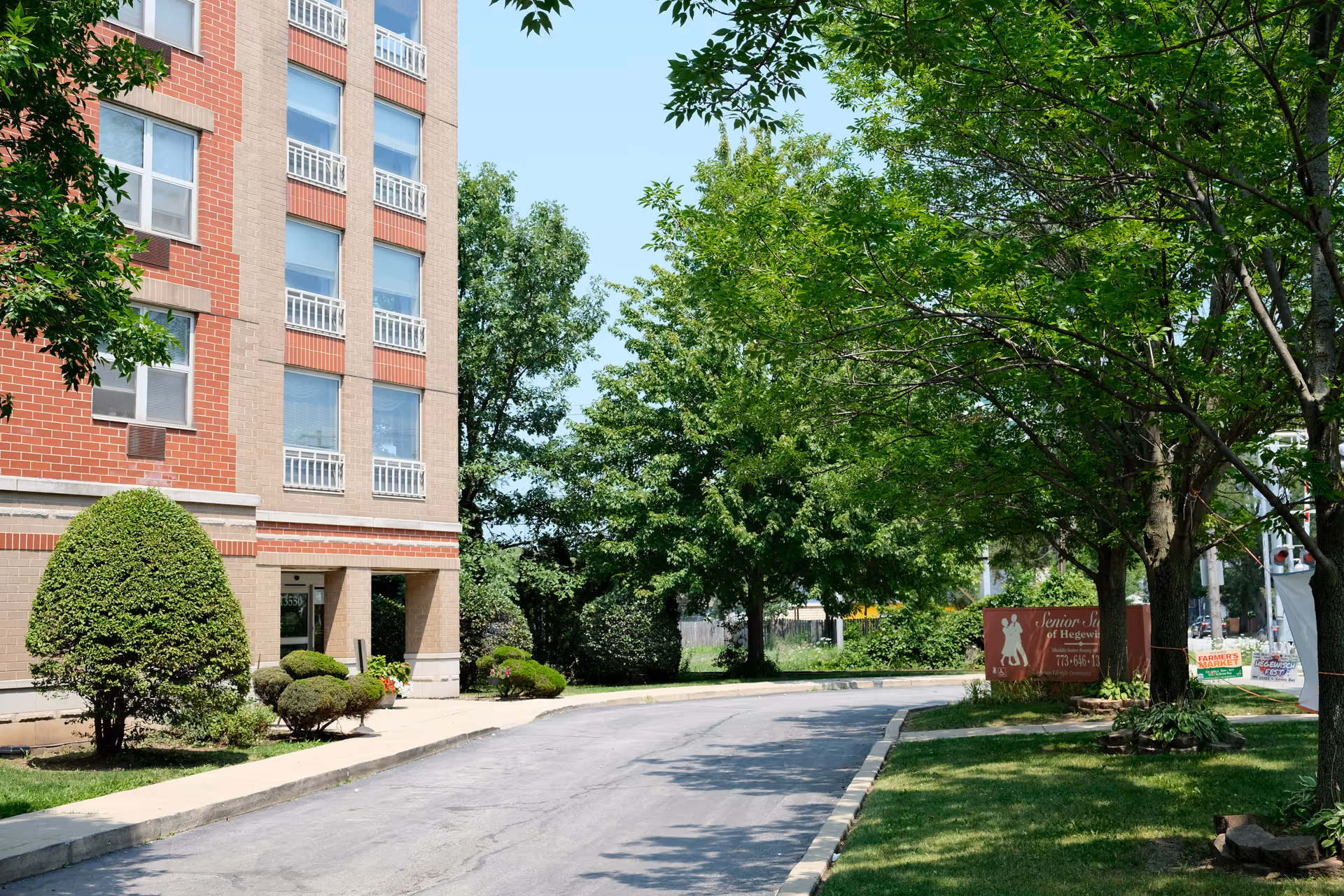 Driveway and landscaped entrance of a brick senior living building with trees and a 'Senior Suites of Hegewisch' sign.