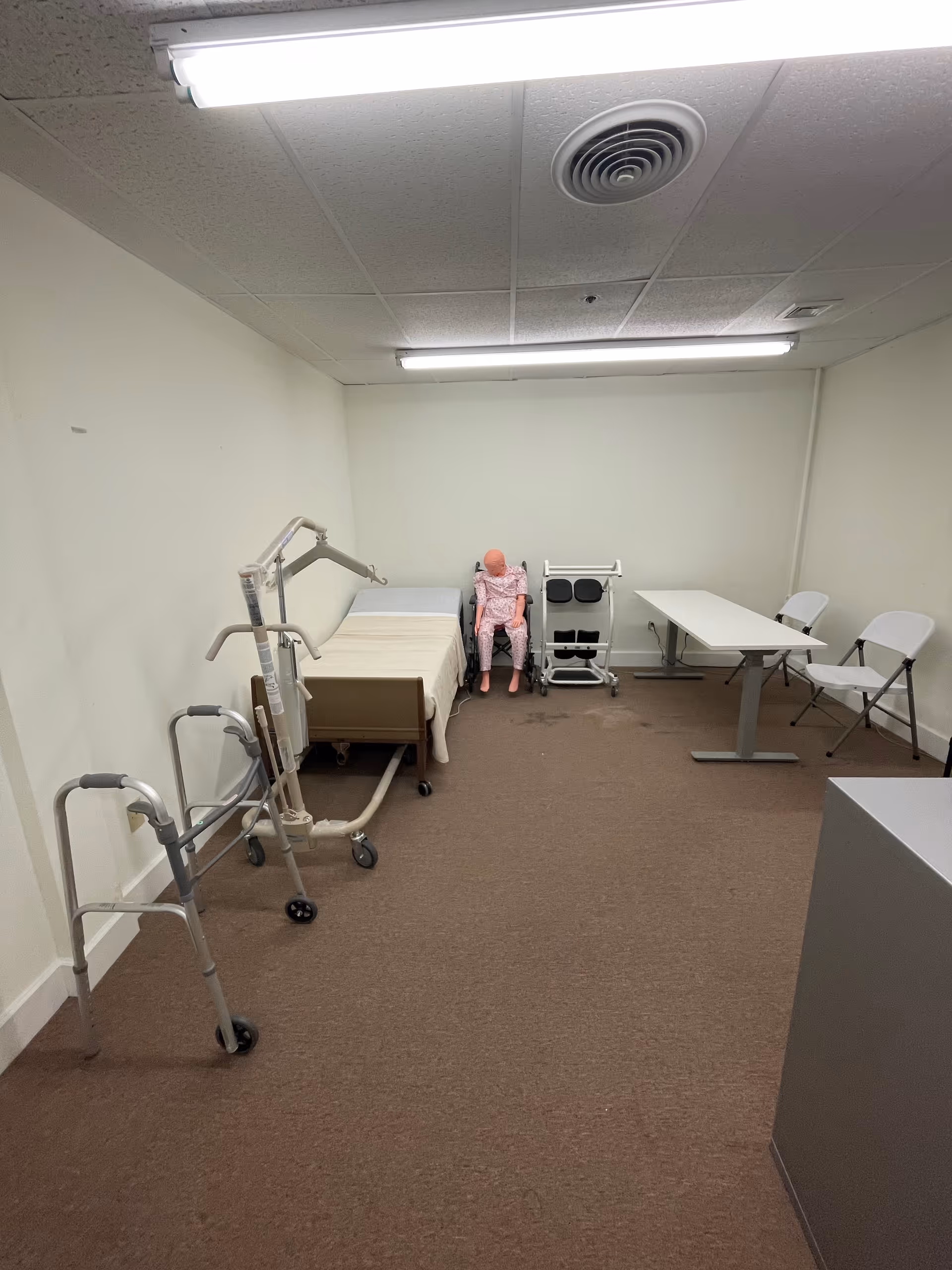Bare clinical room with a bed, mobility aids, a table, and a pink mannequin.