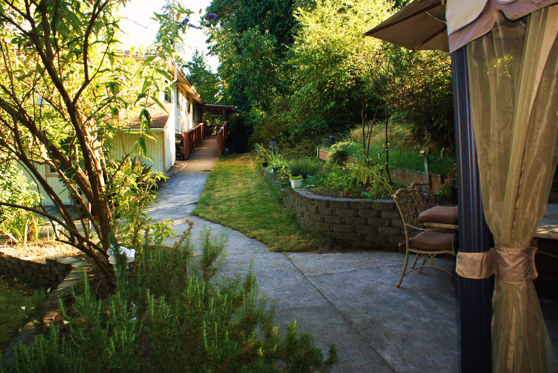 A paved garden path runs alongside a building with a patio and chairs.