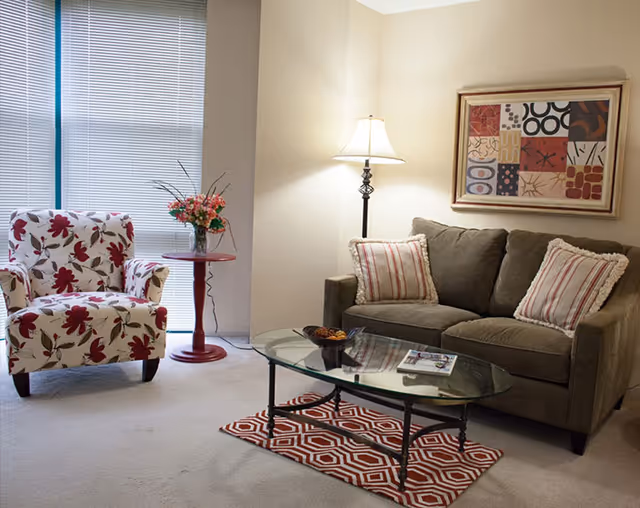 Cozy living room with a floral armchair, gray sofa with striped pillows, glass coffee table on a patterned rug, and a side table with flowers and a floor lamp.