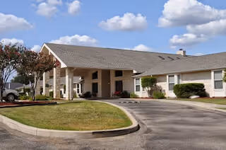 Exterior view of a single-story senior living facility building with a covered entrance, beige siding, and a gray shingled roof under a partly cloudy sky. There is a curved driveway leading to the entrance, with some landscaping including bushes and a tree with red flowers.