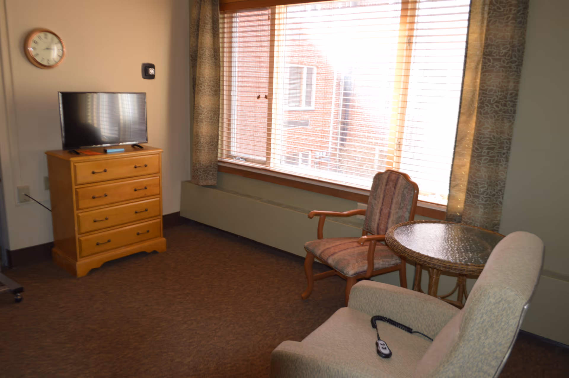 A room with a wooden dresser holding a flat-screen TV, a wall clock, a large window with blinds and patterned curtains, a round glass-top table, a cushioned armchair with a remote control on it, and a wooden chair with armrests.