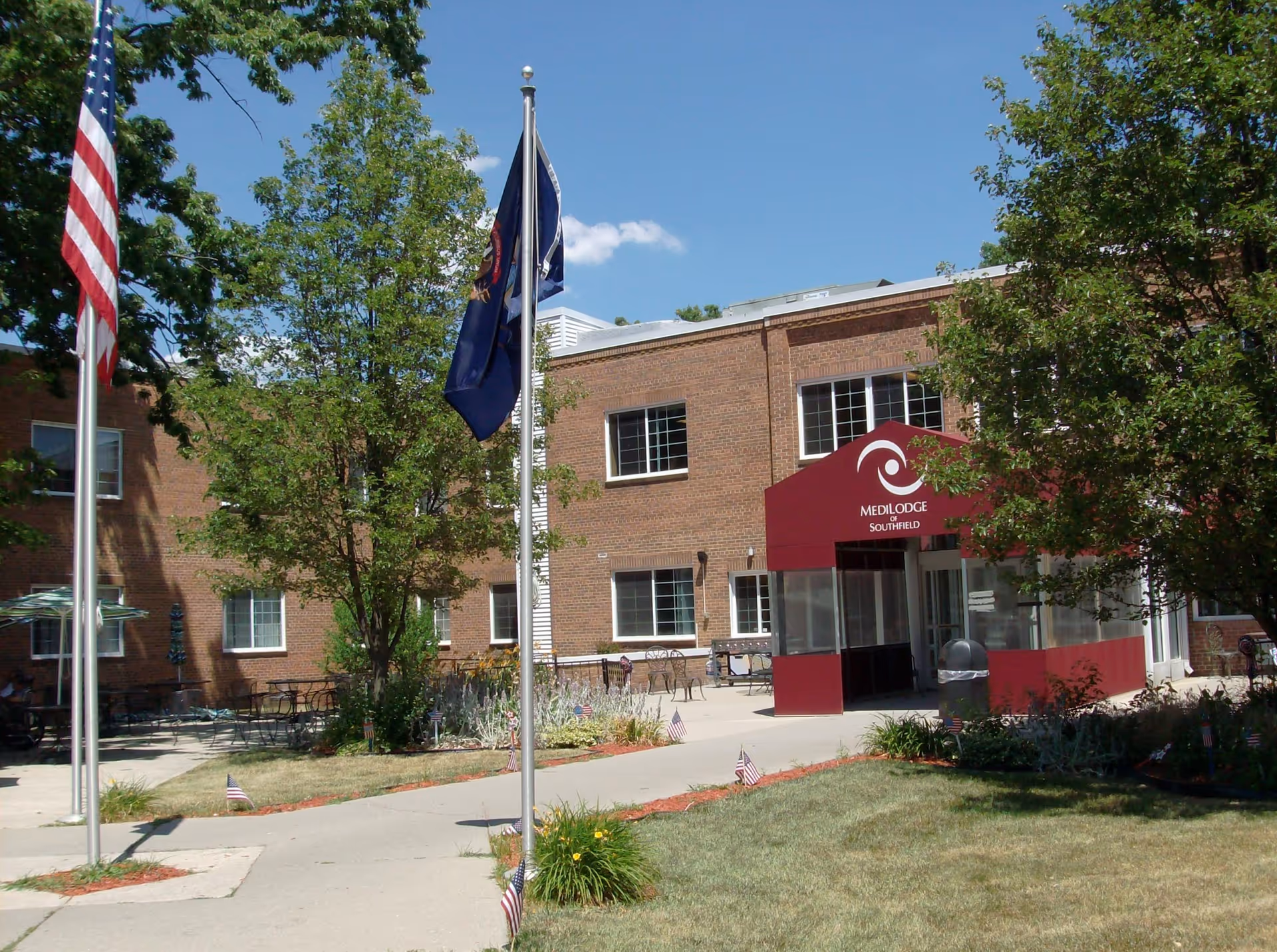 Exterior view of Medilodge of Southfield facility showing a brick building with multiple windows, a maroon entrance canopy with the facility name and logo, two flagpoles with flags, trees, and a walkway surrounded by grass and small American flags.