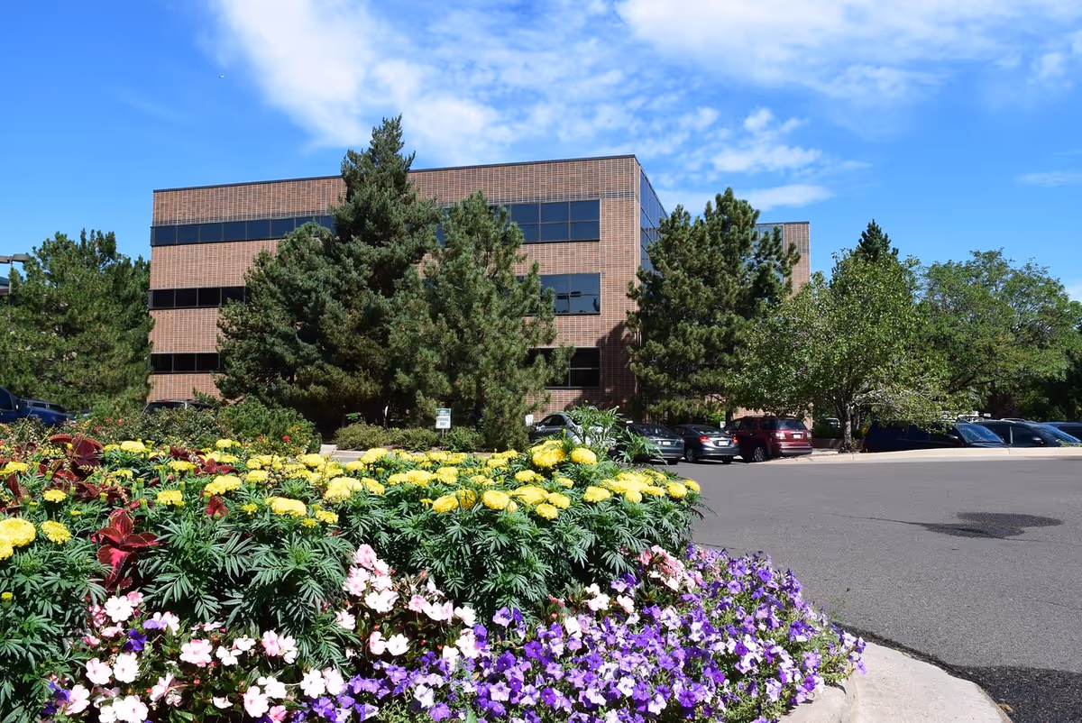 A brick building with large windows, surrounded by trees and colorful flowers in the foreground.