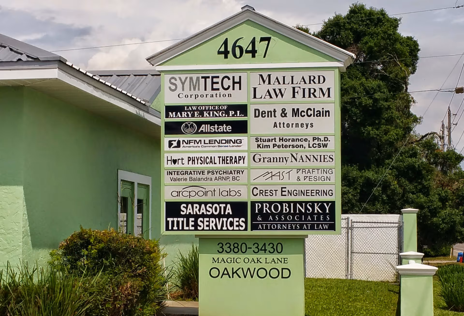 A tall green signage board displays multiple business names in front of a pale green building.