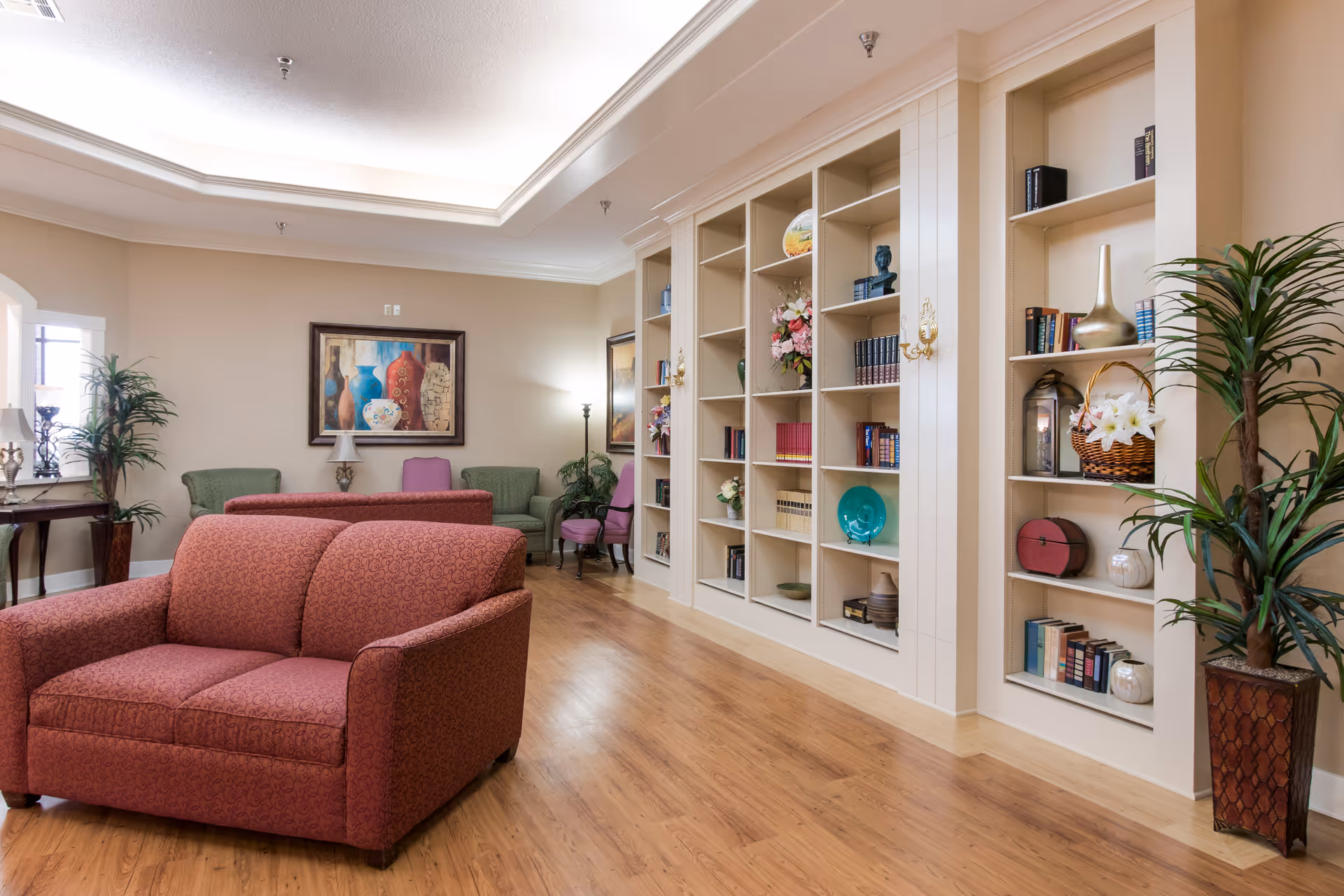 A cozy living room area in a nursing and rehabilitation center featuring a red patterned loveseat in the foreground, green and purple armchairs in the background, built-in white bookshelves filled with books and decorative items, wooden flooring, potted plants, and framed artwork on beige walls.