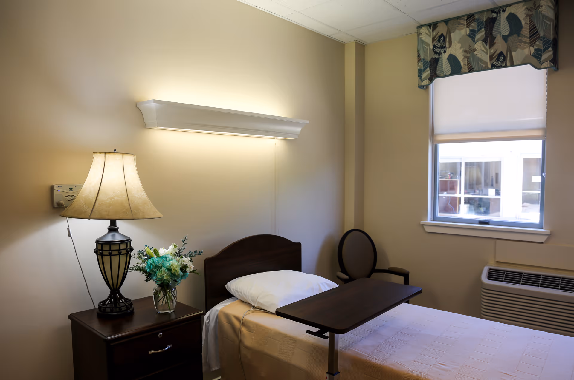 Cozy single bedroom with bed, nightstand, lamp, window, chair, and decorative flowers.