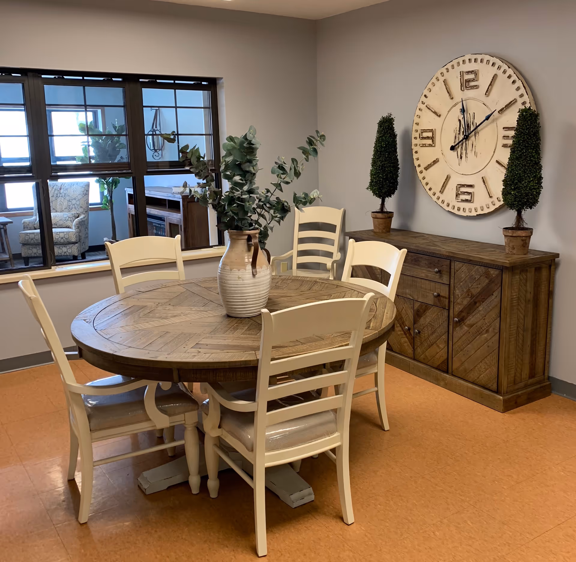 A cozy dining area with a round wooden table surrounded by five cream-colored chairs. A ceramic vase with green foliage is placed in the center of the table. Against the wall is a wooden sideboard with two small potted topiary plants and a large rustic wall clock above it. A window with black framing looks into another room with an armchair and a wooden cabinet.