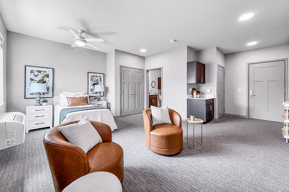 Bright furnished studio suite with bed, seating area, and kitchenette in a modern senior living space.