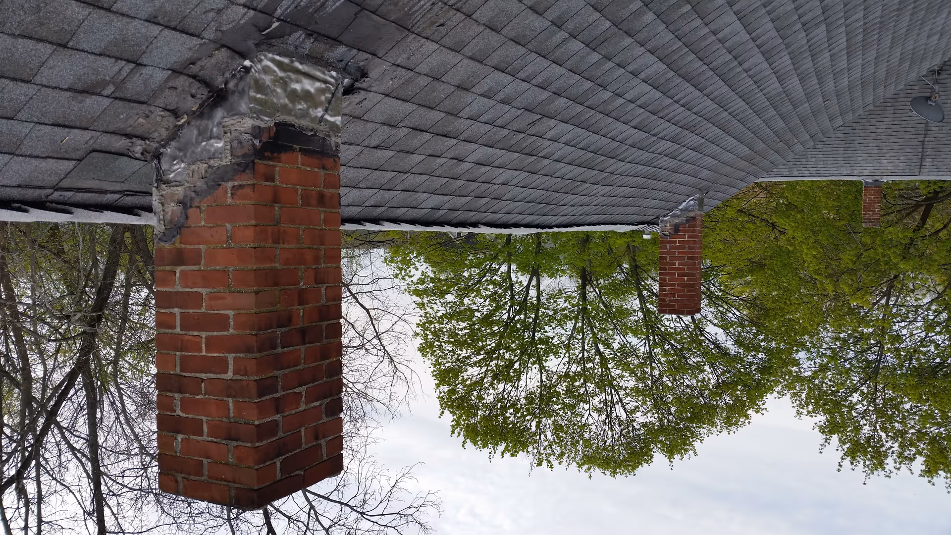 Exterior view of brick columns supporting a shingled roof with leafy trees nearby.