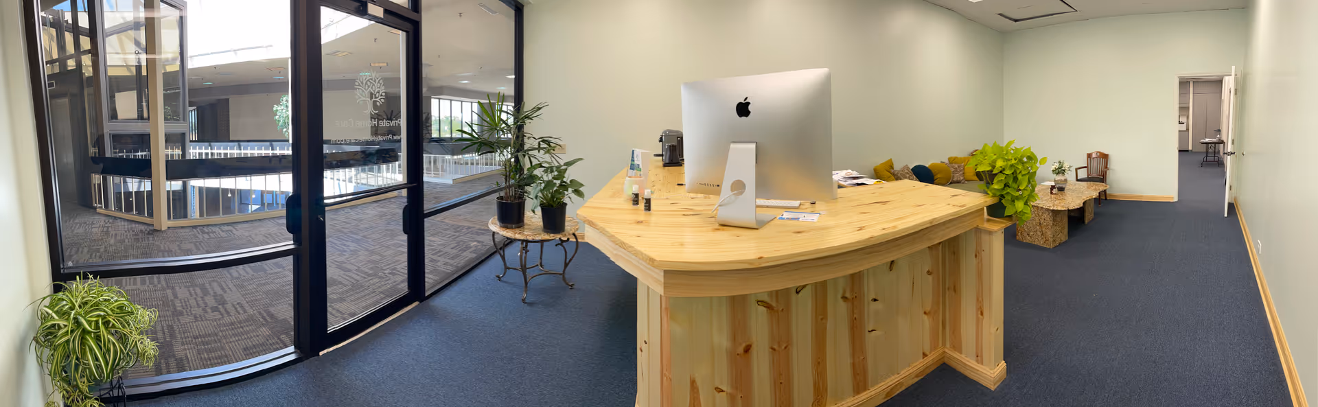 Reception desk with an iMac, plants, and lounge seating in a bright interior lobby.