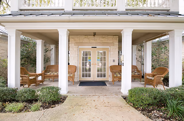 Covered entrance porch with wicker chairs flanking double glass doors and small landscaping in front.