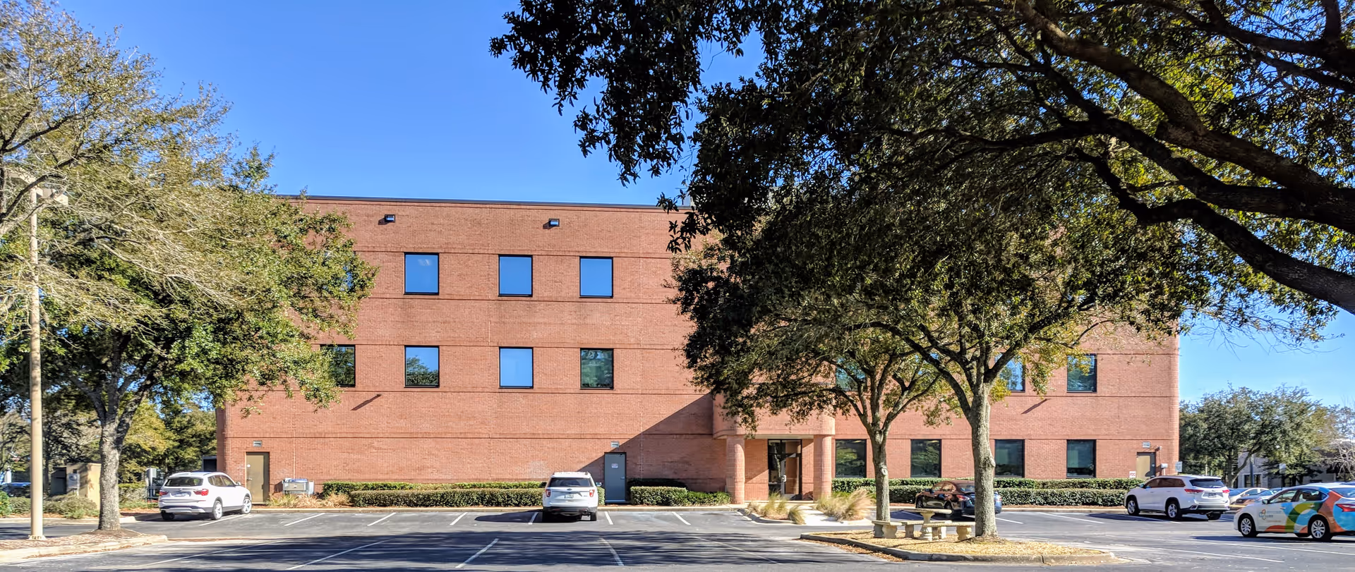 Brick senior living facility with a parking lot, trees, and a clear blue sky.