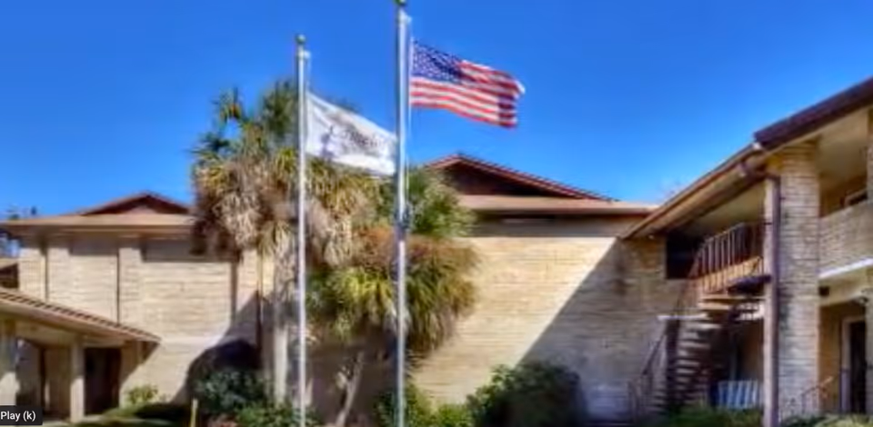 Exterior view of Camelot Chateau Assisted Living with flags, brick walls, and palm trees.